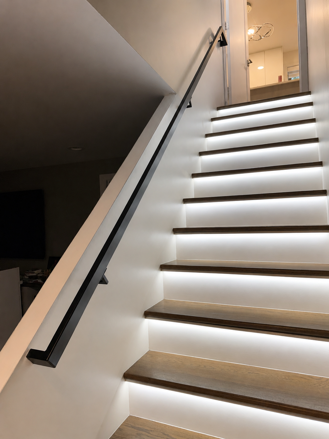 Interior view of a staircase with wood steps and integrated LED lighting. A black metal handrail runs along the wall, with a door and part of a ceiling light fixture visible at the top.