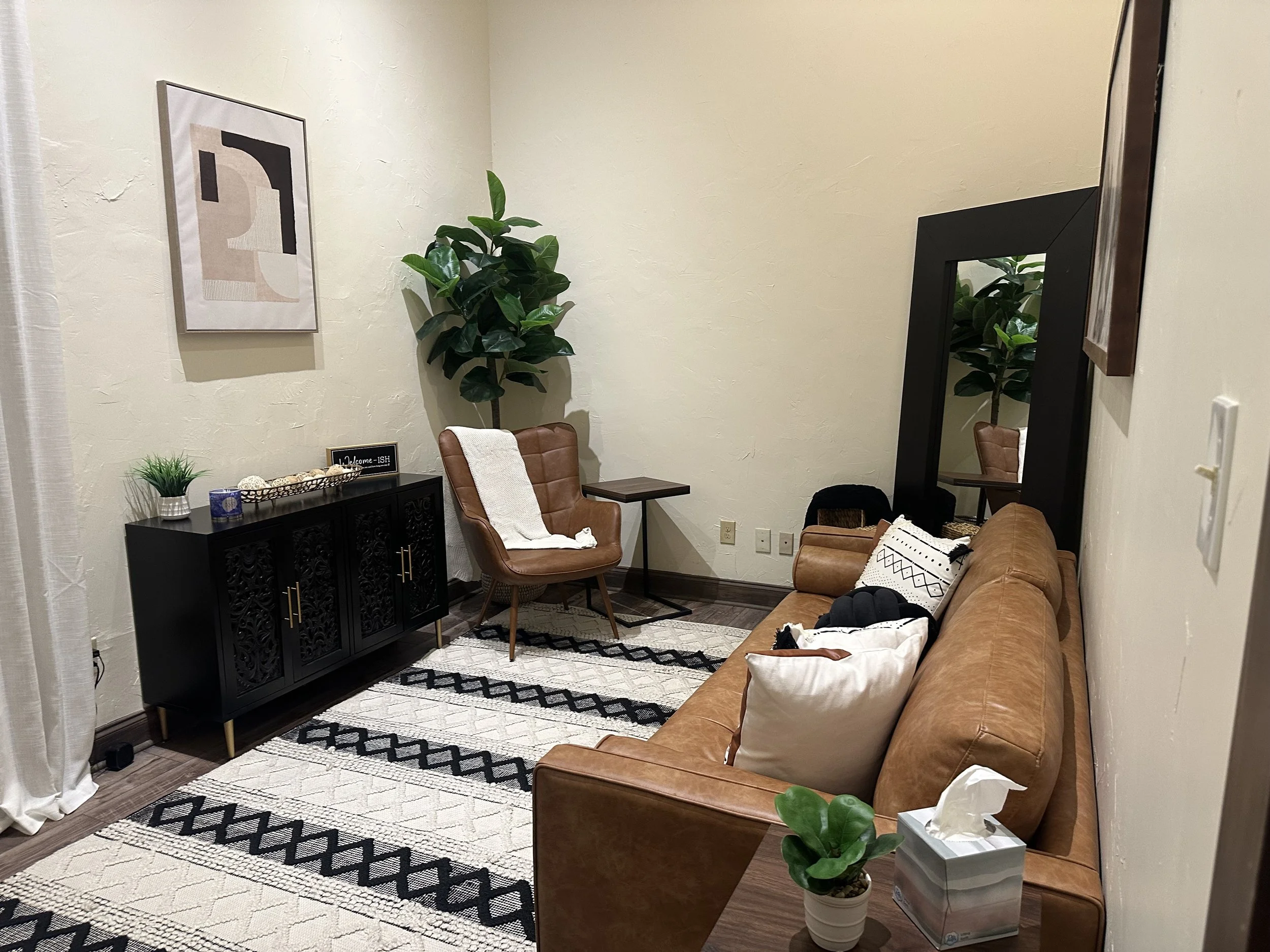 Living room with tan leather sofa, patterned rug, black sideboard, brown accent chair, large plant, framed artwork, mirror, and decorative items.