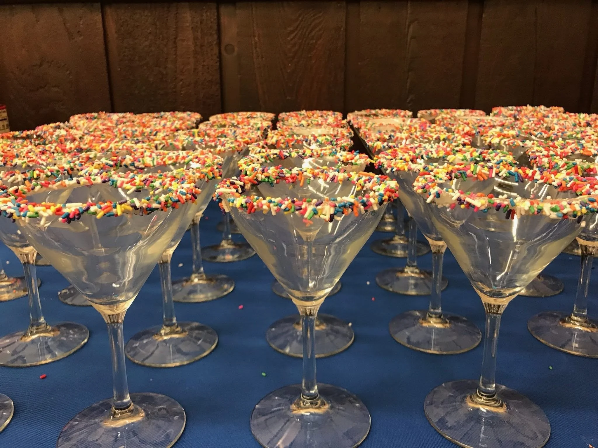 Multiple cocktail glasses with a sugar and rainbow sprinkles rim, arranged on a blue tablecloth against a wooden wall background.
