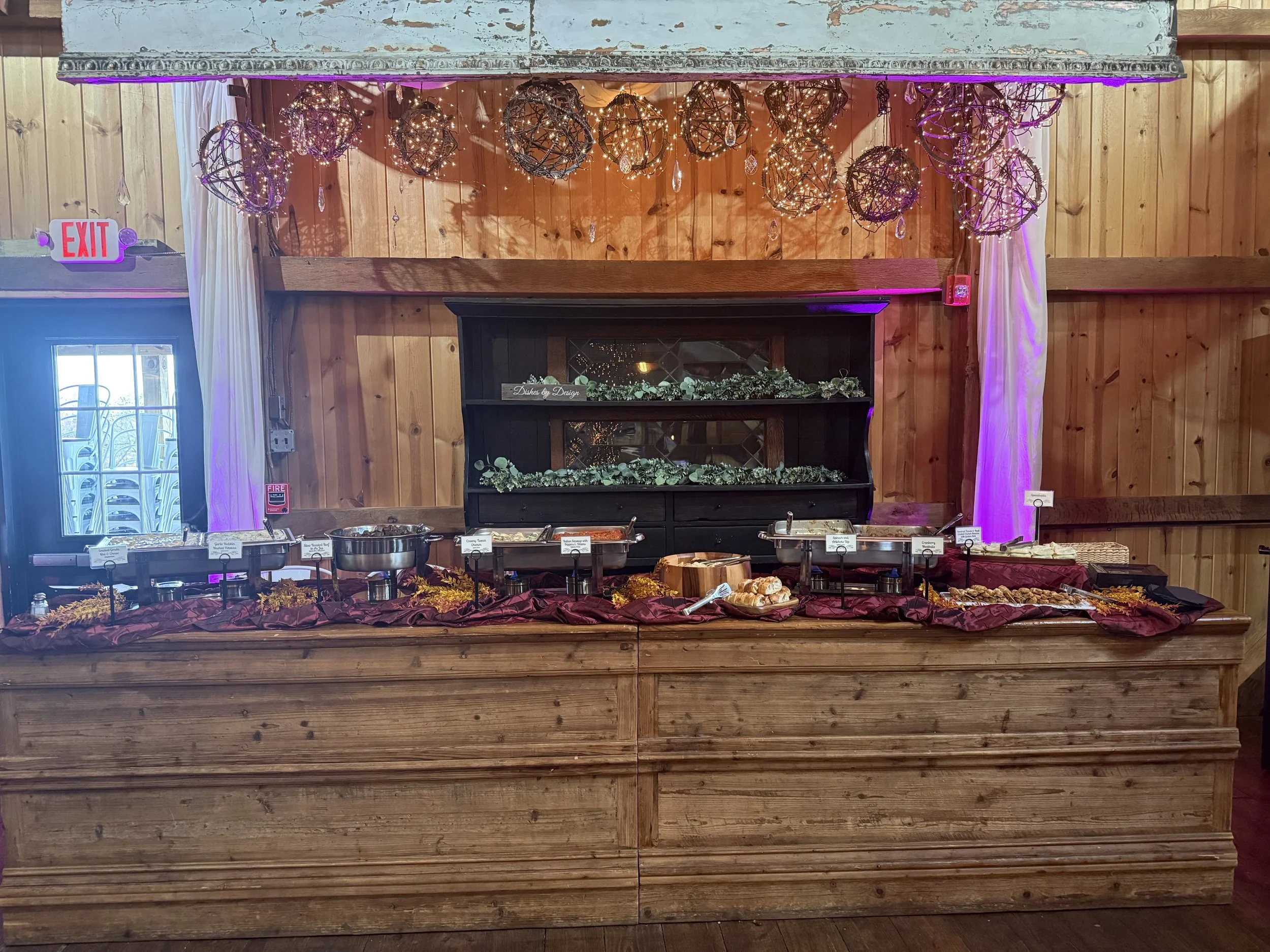 Buffet table with chafing dishes and food in a wooden restaurant or event space, decorated with string lights and hanging spherical decorations, with a wooden wall backdrop and purple uplighting.