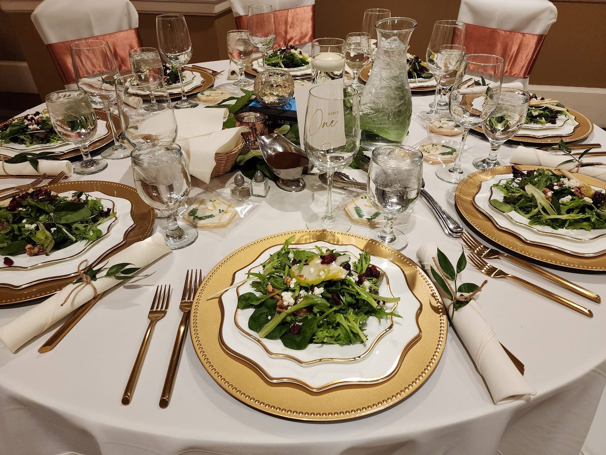 Table setting with salad plates, gold-rimmed china, silverware, multiple glasses of water, a pitcher of water, and decorative greenery on a white tablecloth for a formal dinner.