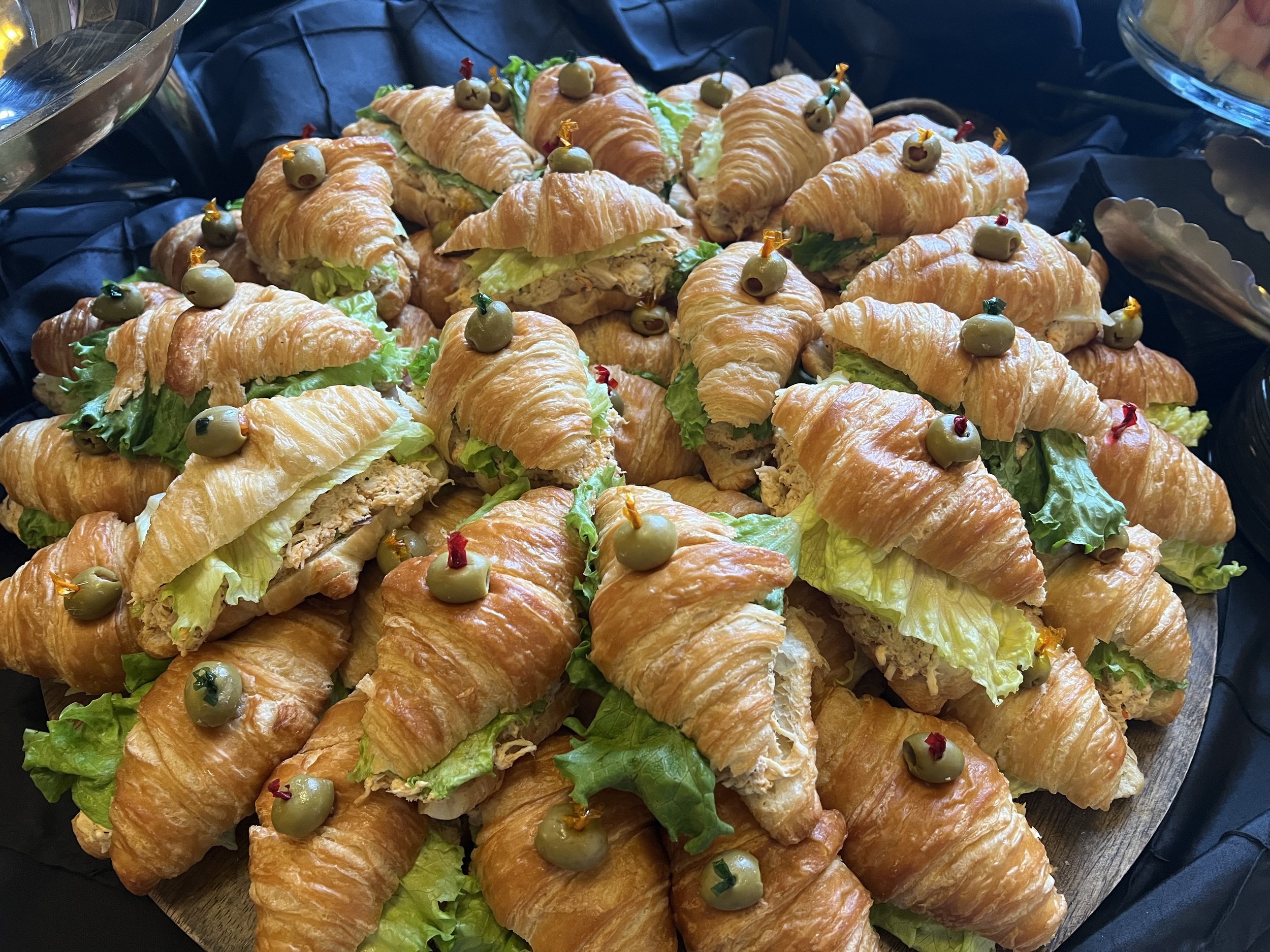 A platter of mini croissant sandwiches filled with lettuce and chicken salad, each topped with a green olive and a small decorative pick.