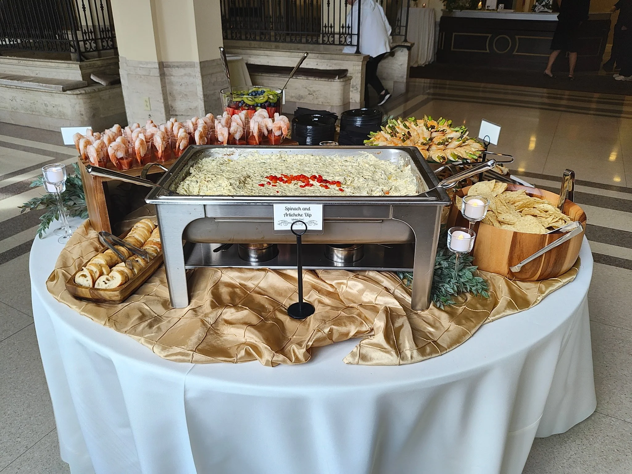 Buffet table with shrimp, spinach and artichoke dip, shrimp, toast points, and crackers decorated with candle holders and greenery in an event hall.