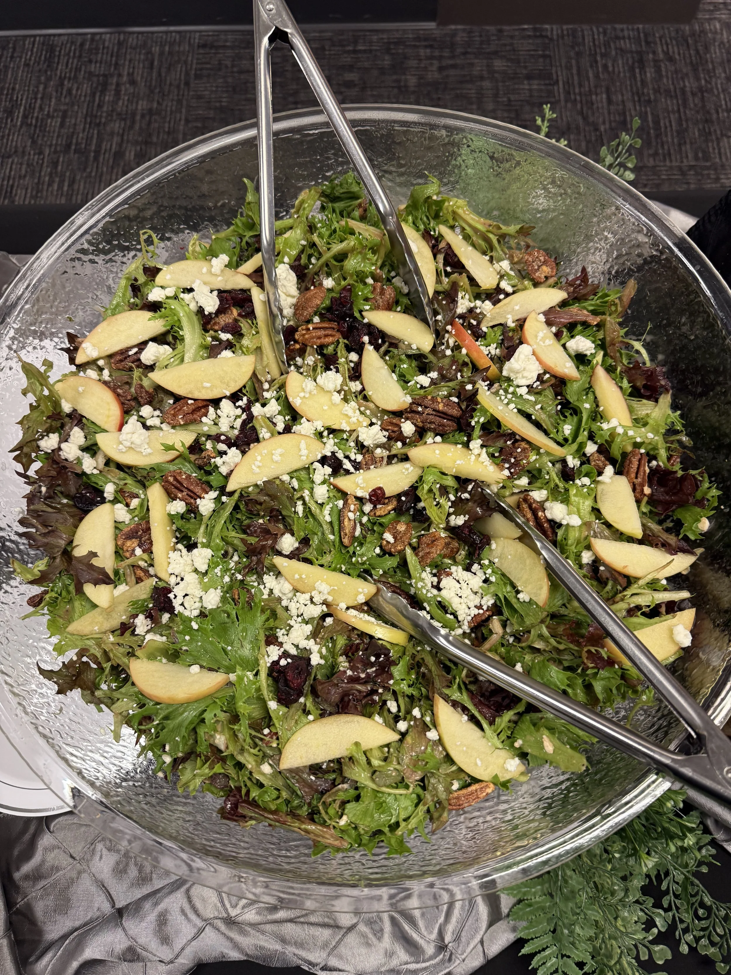 A bowl of mixed green salad with sliced pears, crumbled cheese, pecans, and dried cranberries, with tongs resting on top.