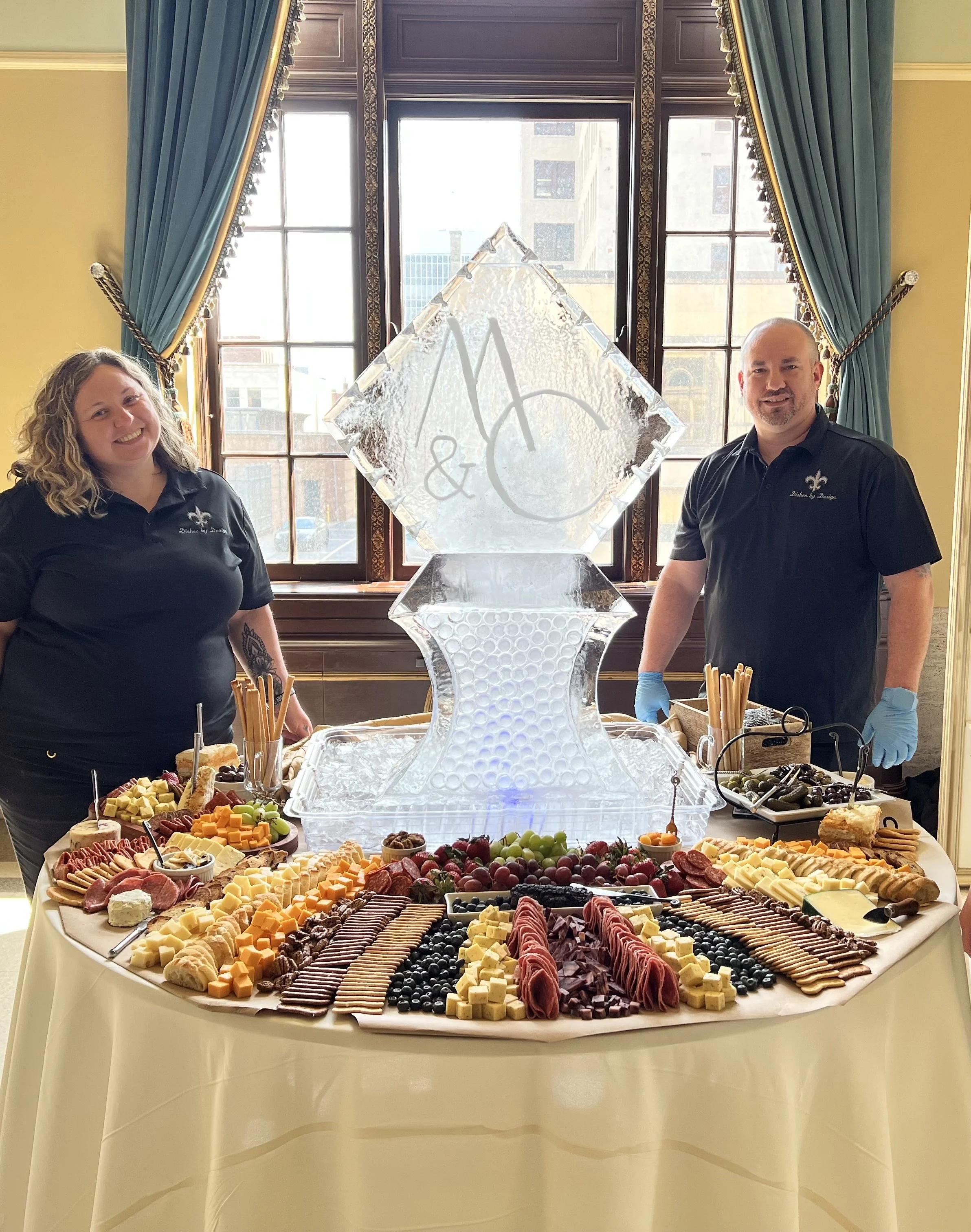 Two catering staff behind a table with a large ice sculpture centerpiece, surrounded by a variety of cheeses, meats, fruits, olives, and crackers in a formal room with large windows and blue curtains.