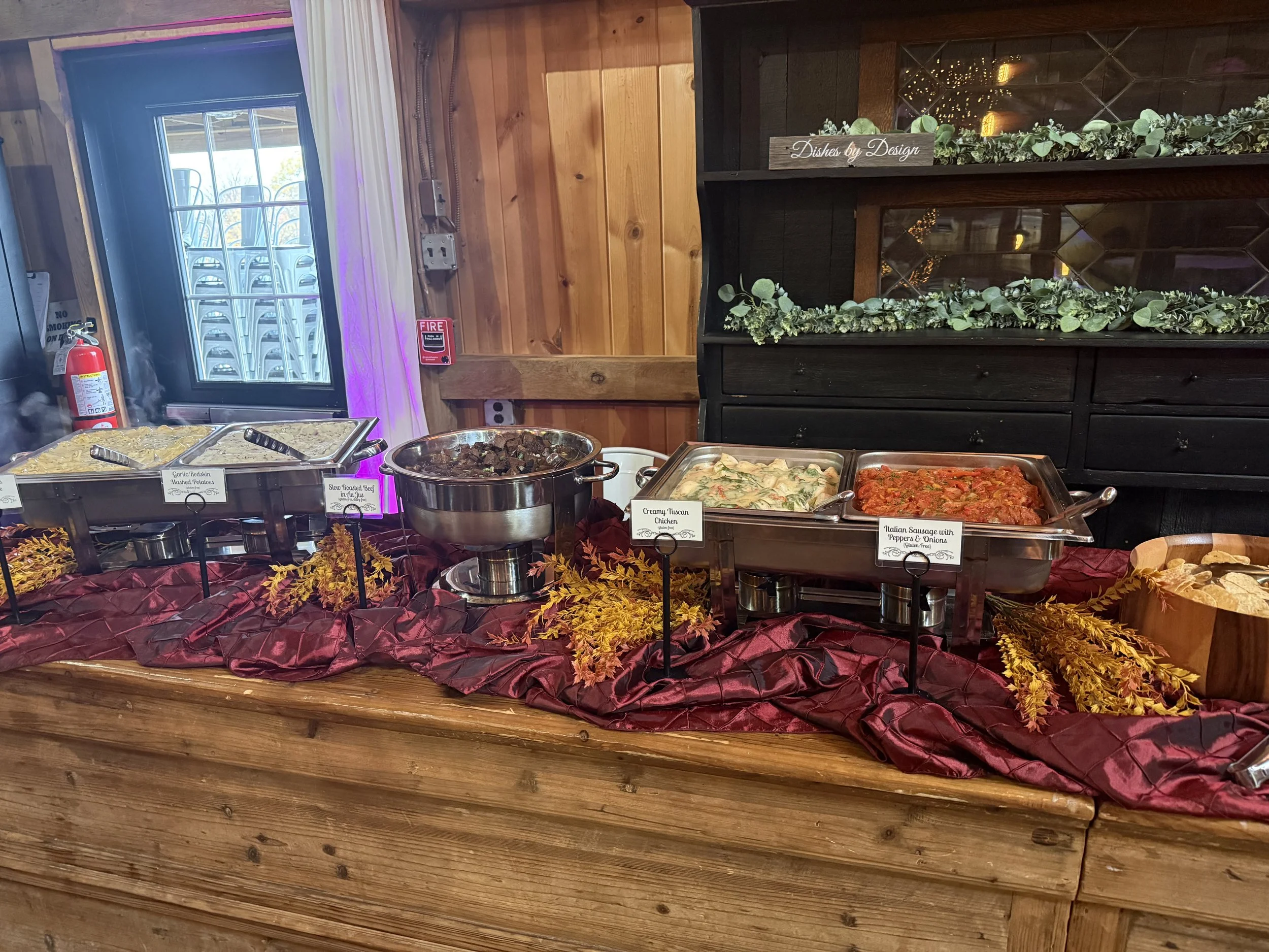 A buffet table with various dishes, including garlic mashed potatoes, slow roasted beef, creamy Tuscan chicken, and Italian sausage with peppers and onions, decorated with autumn leaves.