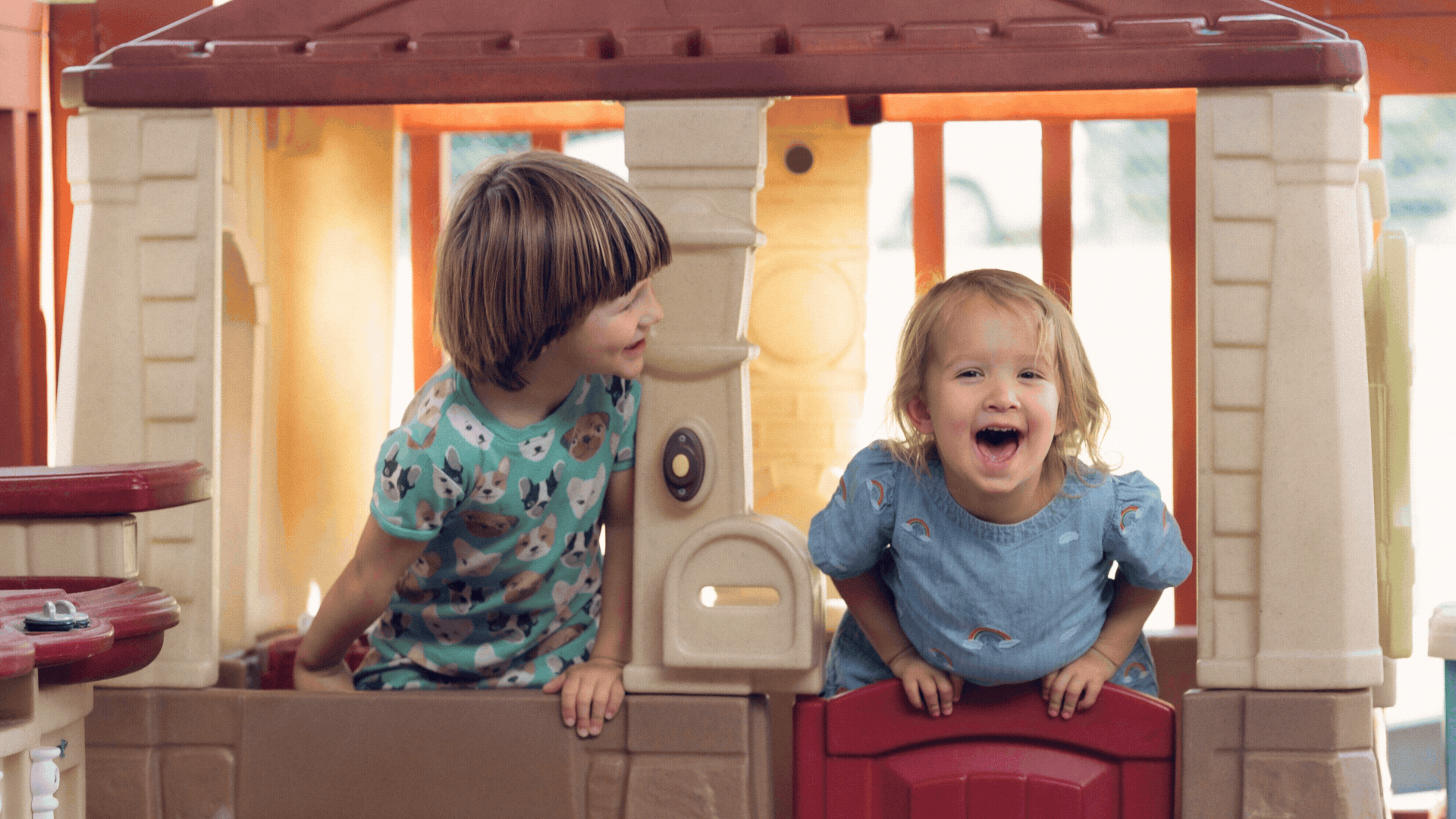 Two young children, a boy and a girl, playing and smiling inside a plastic playhouse with a brown roof and beige walls.