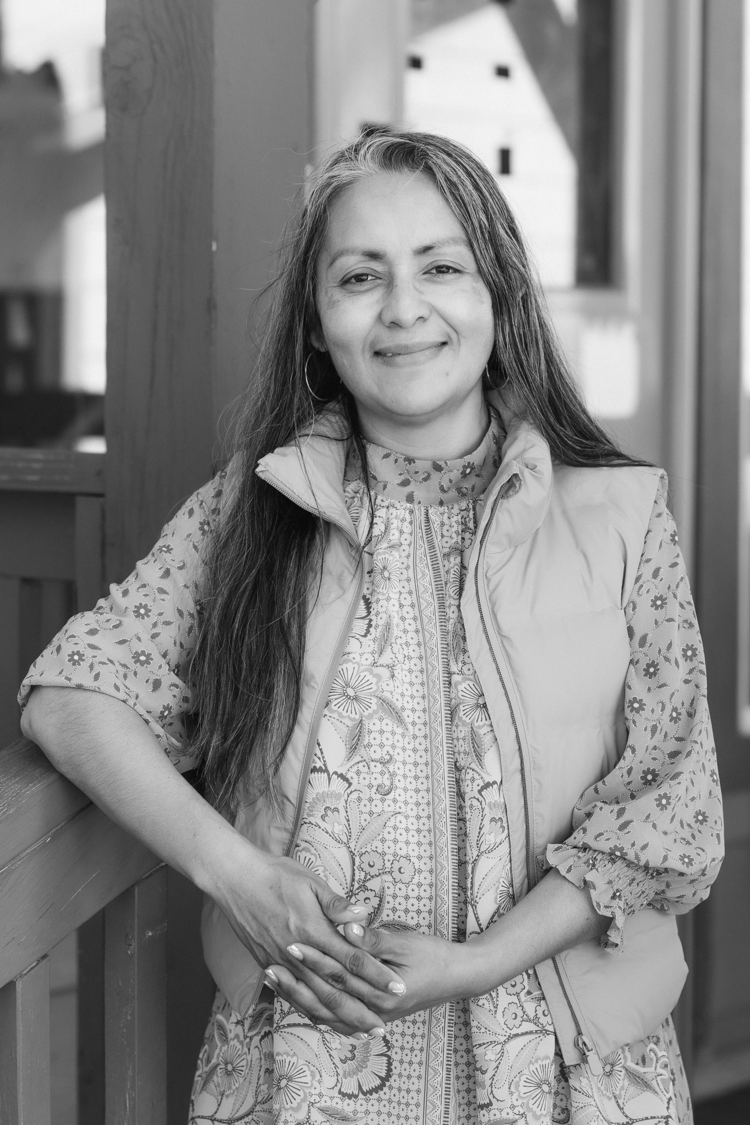 A woman with long hair and hoop earrings standing outdoors and smiling, leaning on a wooden railing, wearing a patterned dress and a light-colored vest.