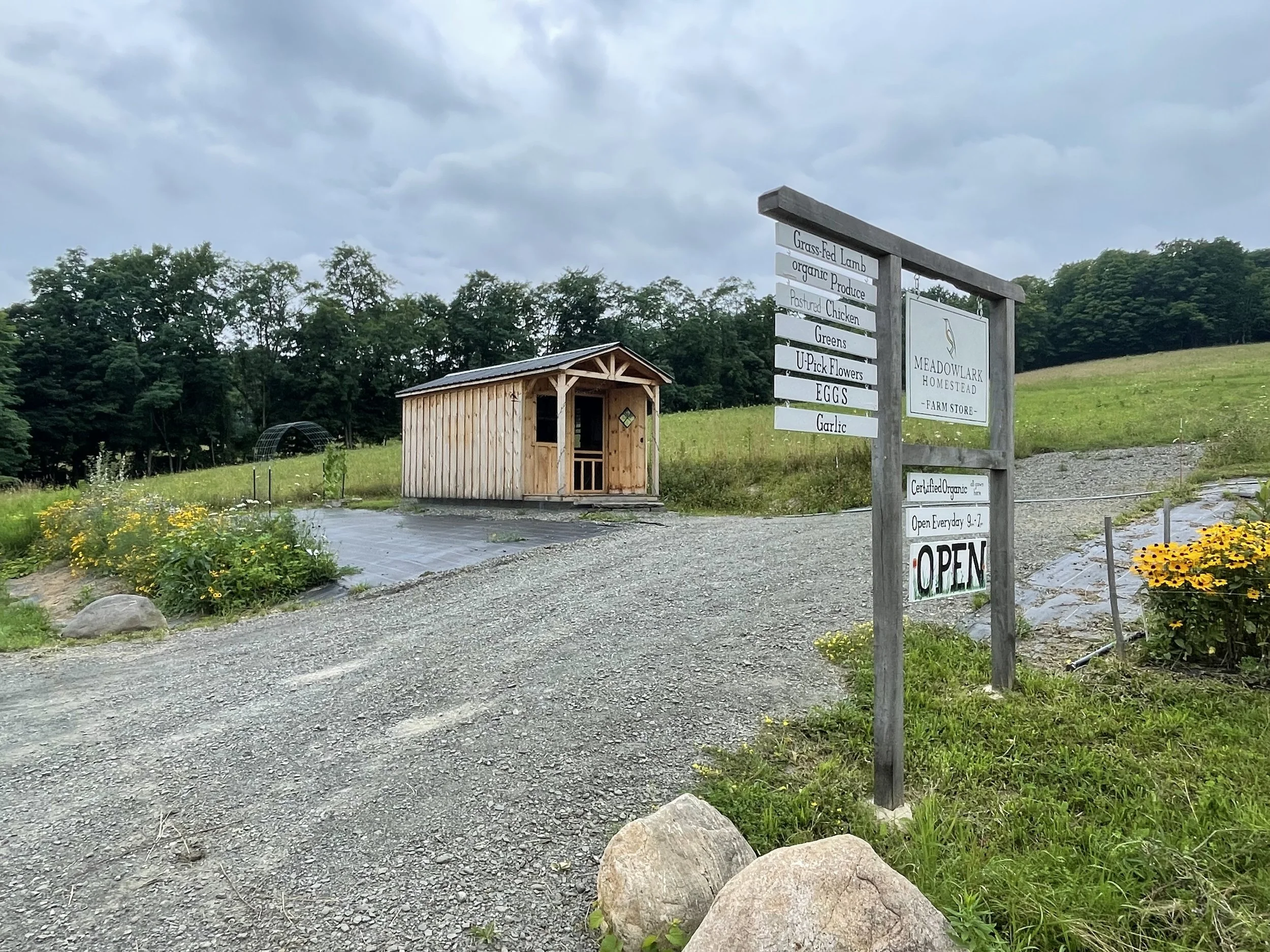 A farm store sign with various items listed, next to a small wooden shed, surrounded by greenery and flowers.
