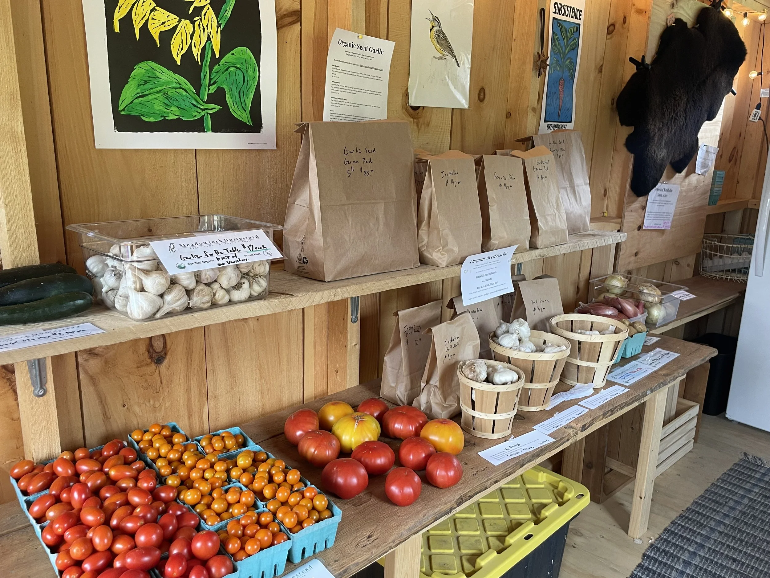 Display of fresh vegetables at a farm stand, including cherry tomatoes, heirloom tomatoes, garlic, zucchini, onions, and paper bags of various produce against a wooden wall decorated with posters and art.