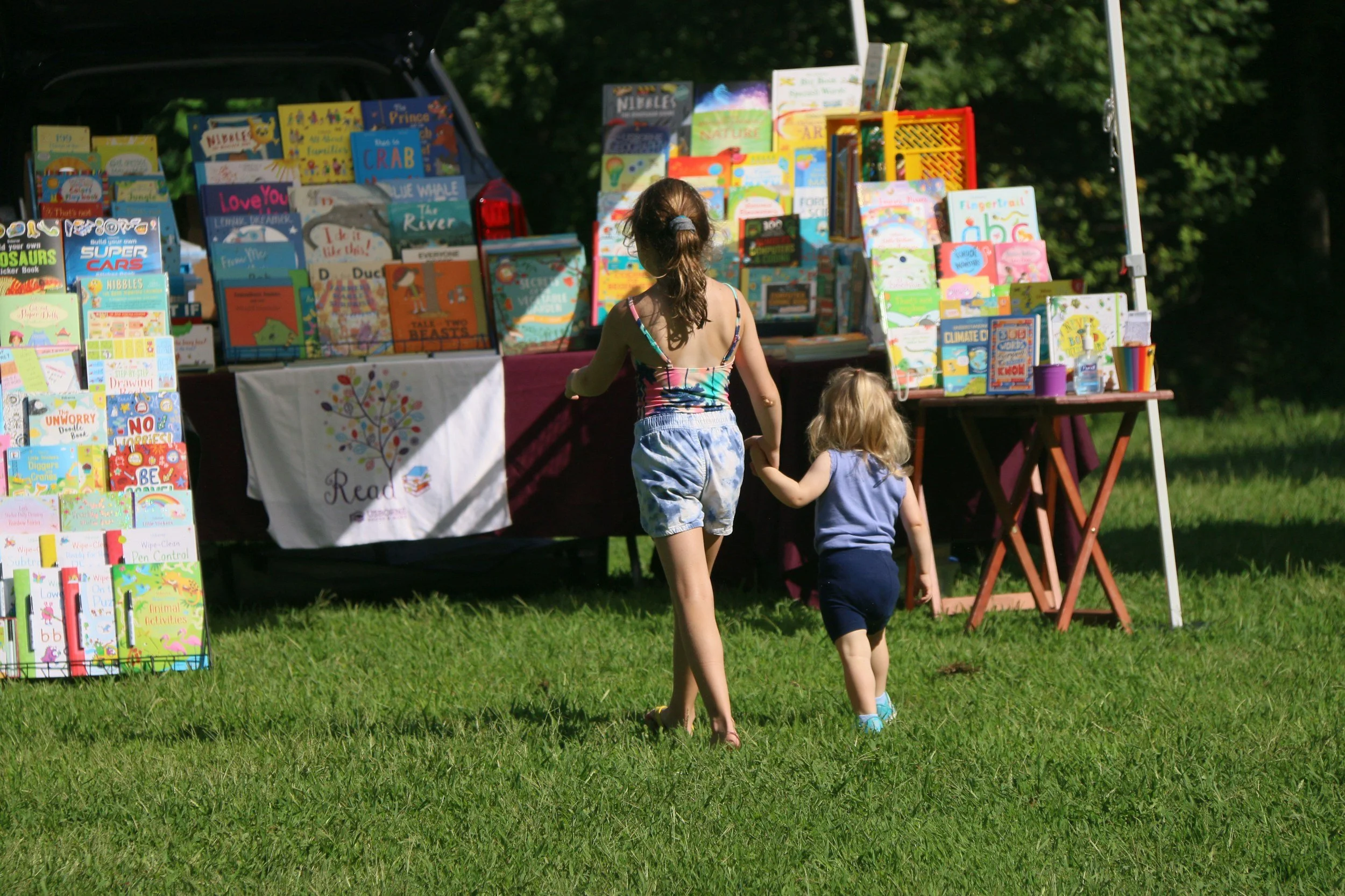 A girl and a young child walking past a book booth at an outdoor fair or market, with children’s books displayed on tables and racks.