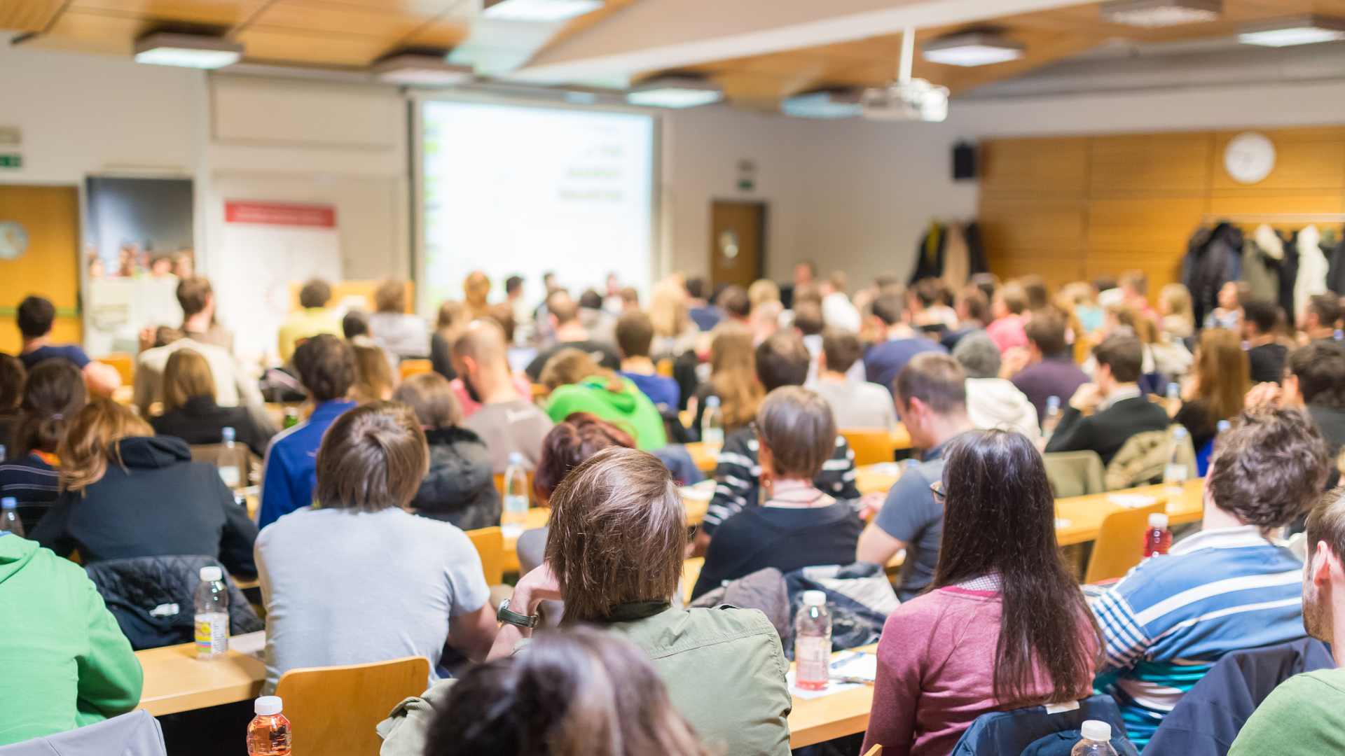 A large group of people attending a conference or lecture in a classroom or lecture hall with a presentation screen at the front.