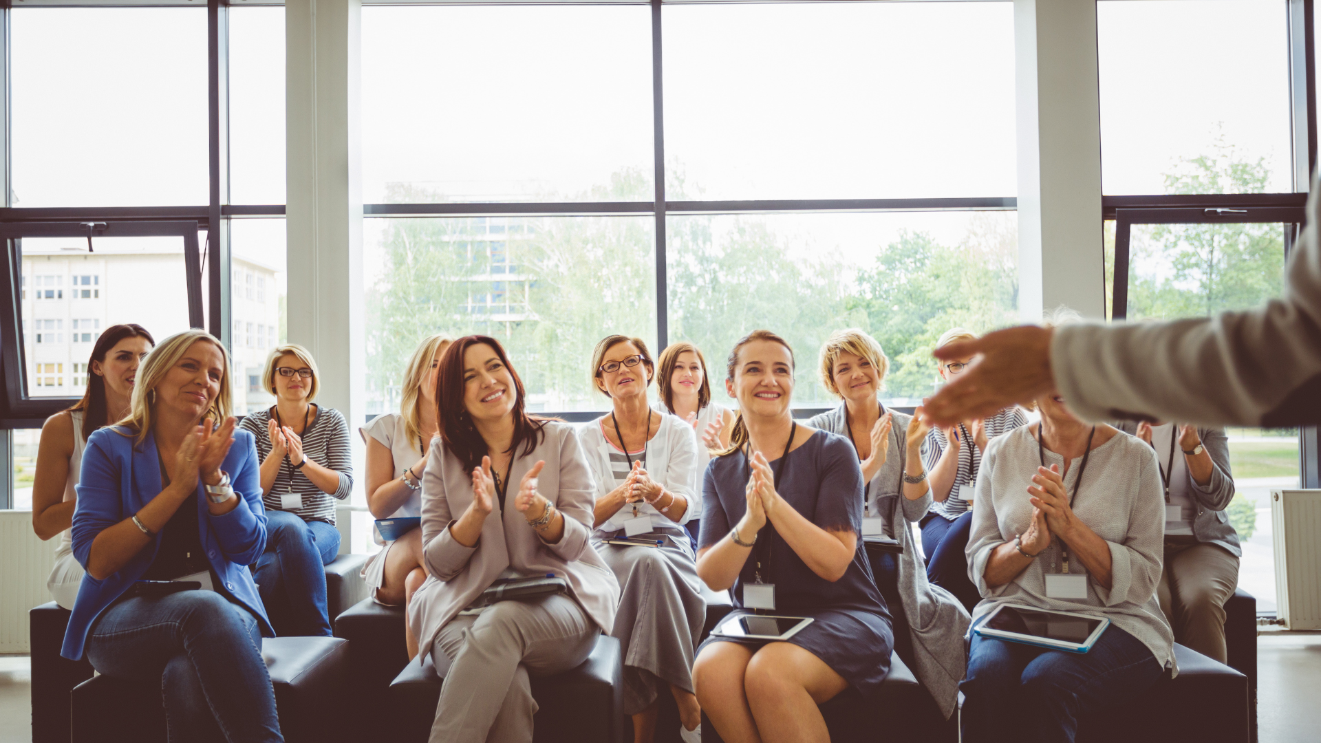 Group of women at a business seminar clapping and smiling while listening to a presenter in a bright room with large windows.