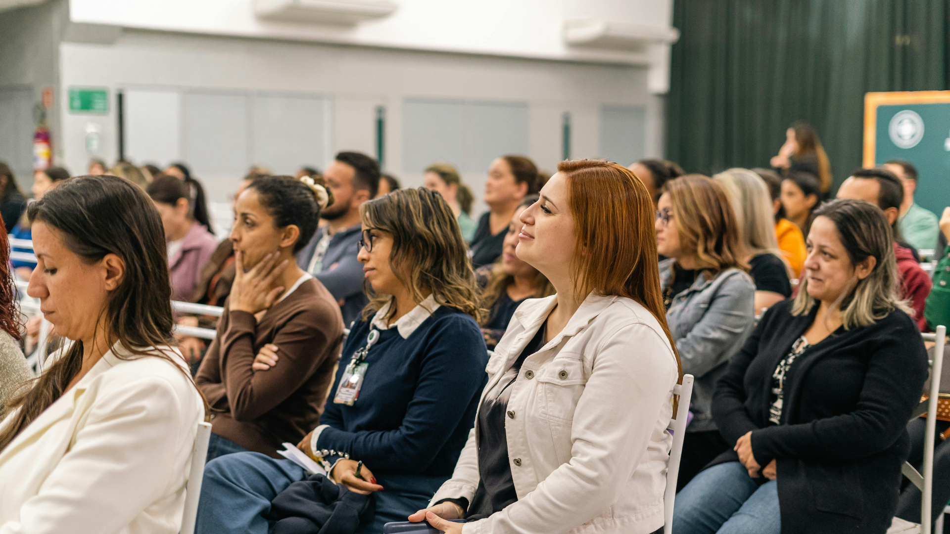 Audience members seated in rows at a conference or seminar, attentively listening to a speaker, in a large indoor venue.