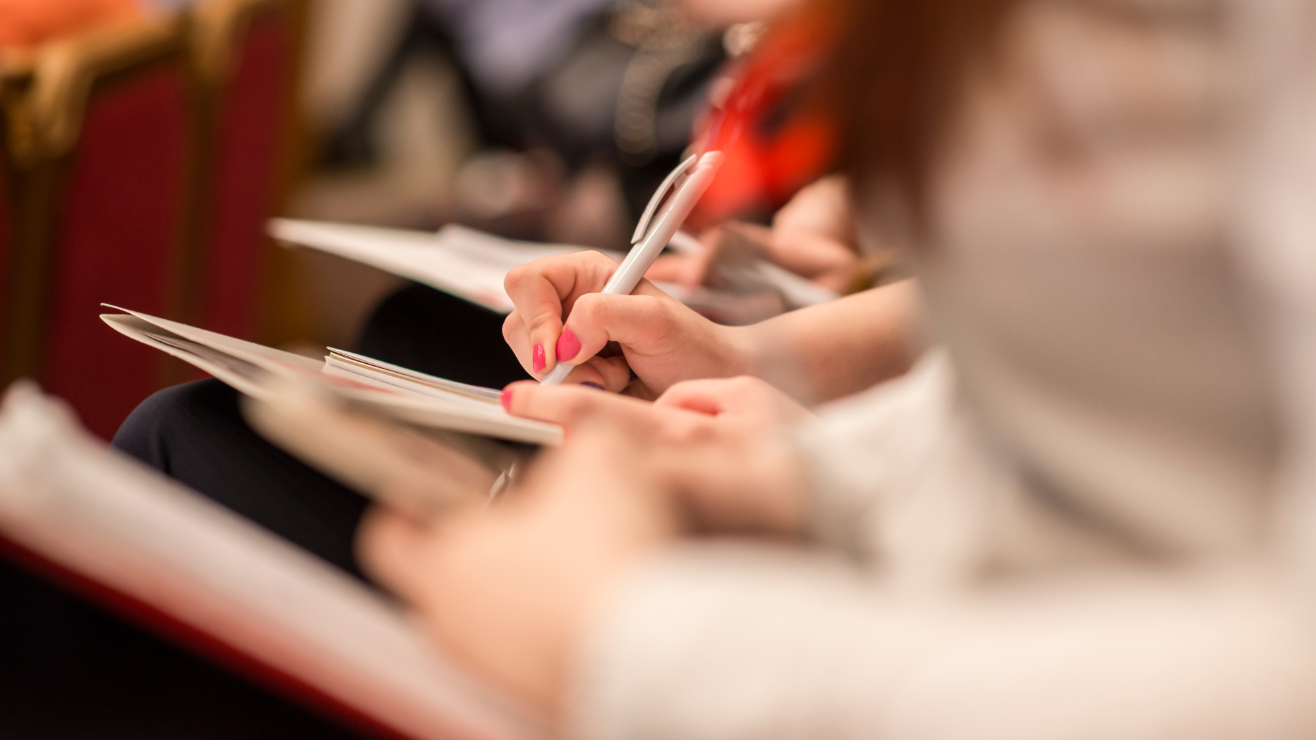 Close-up of a person taking notes with a pen on paper during a meeting or conference, with other attendees in the background.