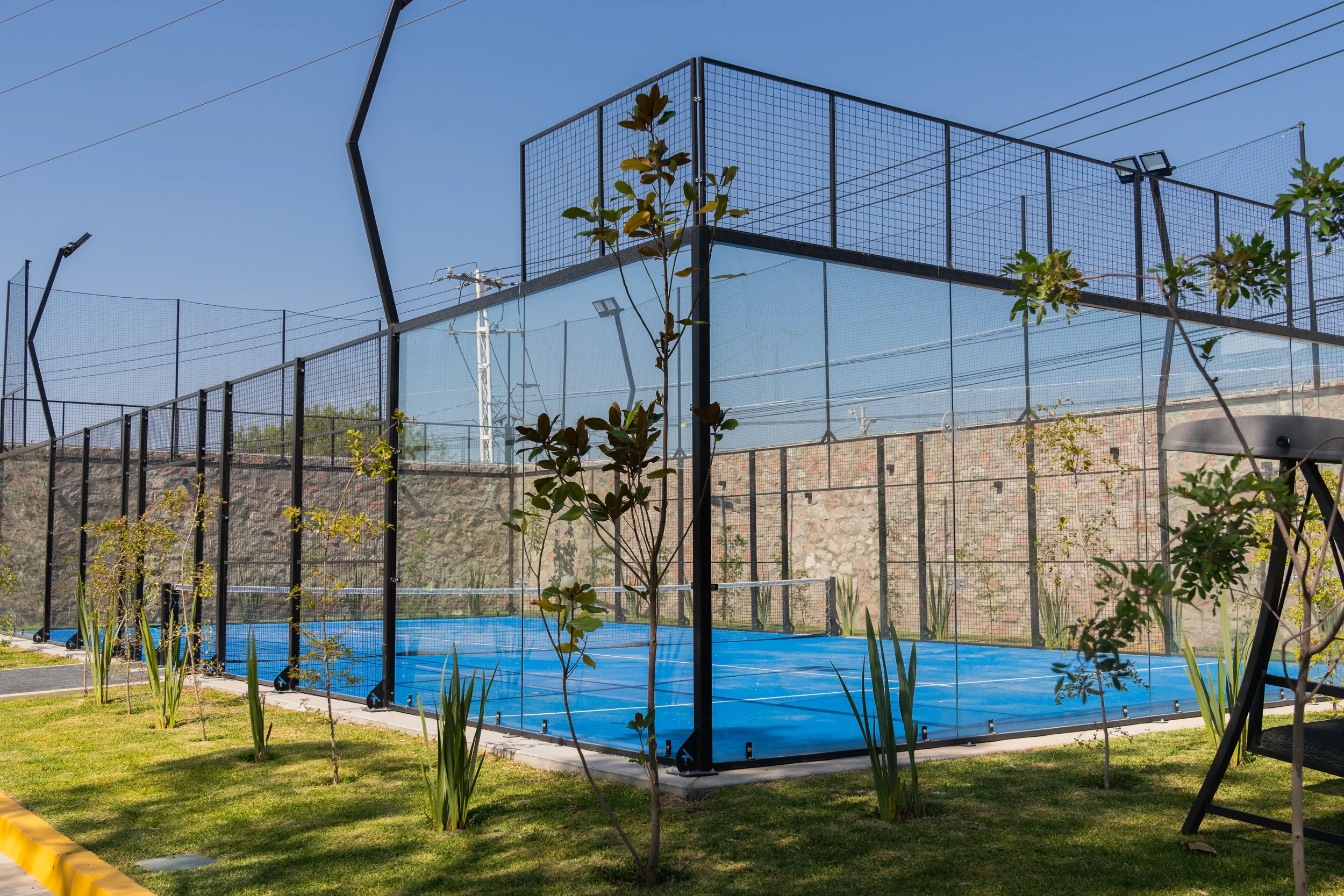 A tennis court with a blue surface enclosed by black and mesh fencing, surrounded by small trees and plants, under a clear blue sky.