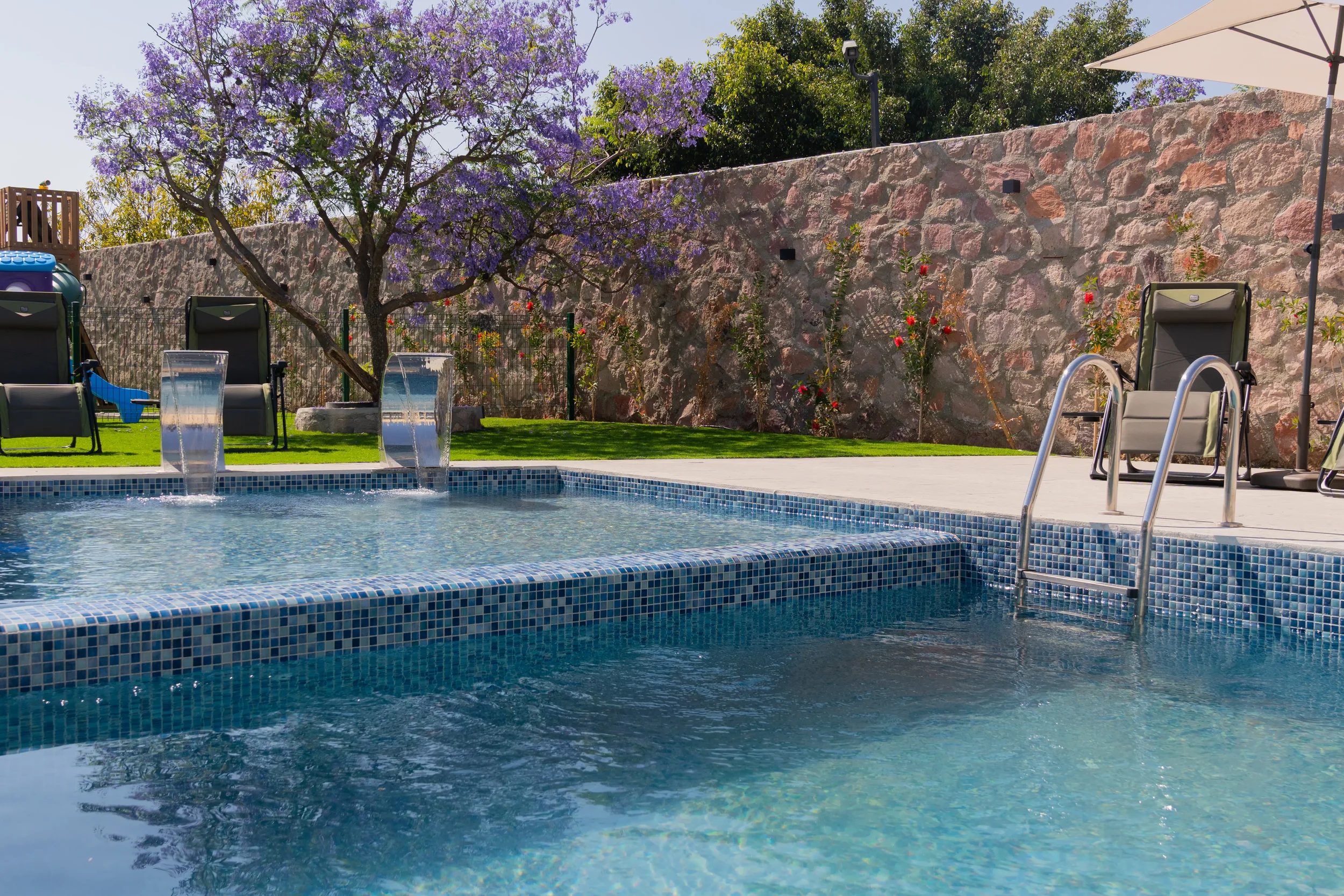 A backyard swimming pool with waterfalls, surrounded by lounge chairs, a stone wall, and a flowering tree with purple blossoms.