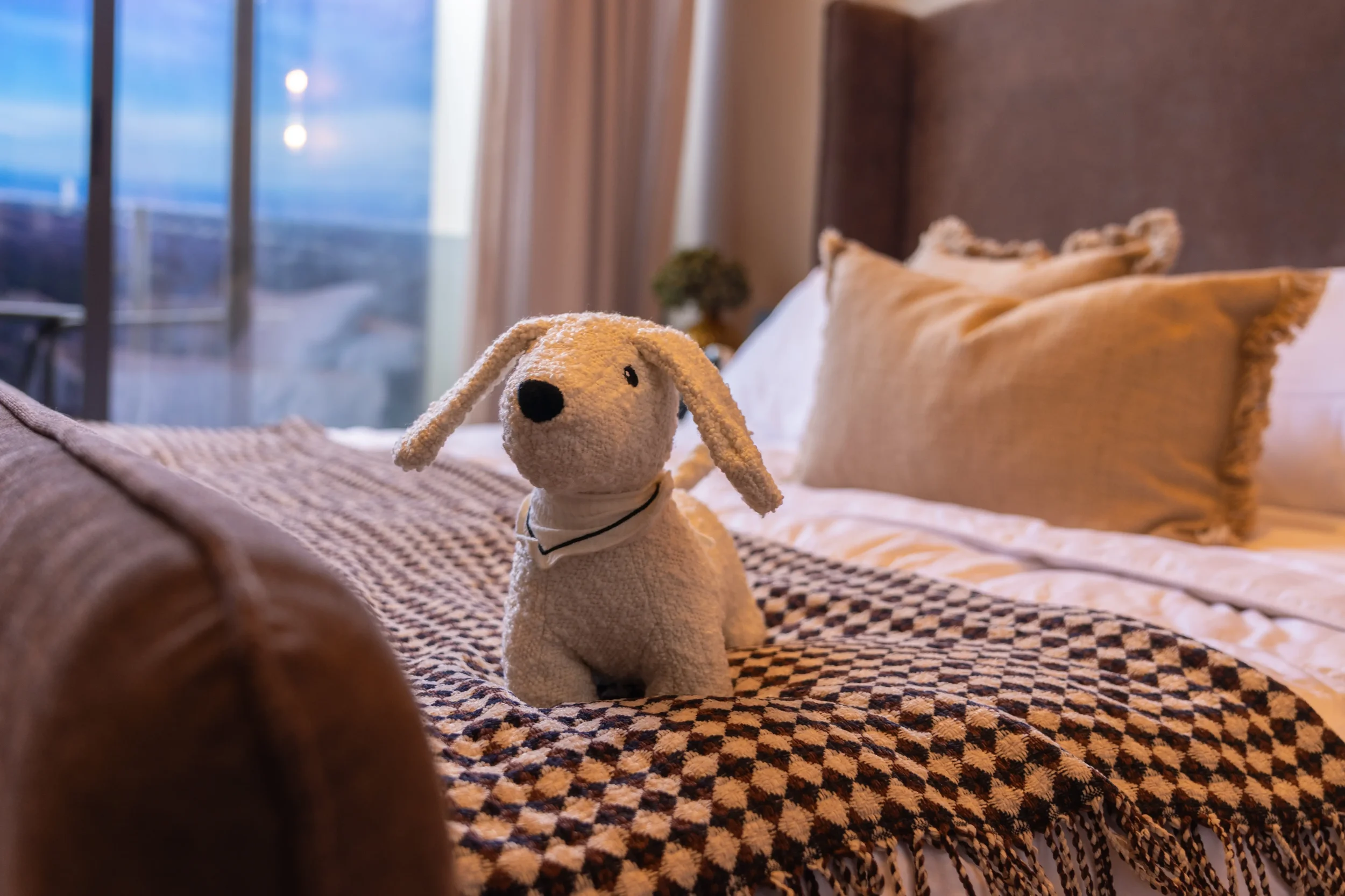 A plush white dog stuffed animal with floppy ears and a black nose sitting on a bed with a brown and black patterned blanket in a cozy bedroom. In the background, there is a sliding glass door with curtains, and a night or early morning sky visible outside.