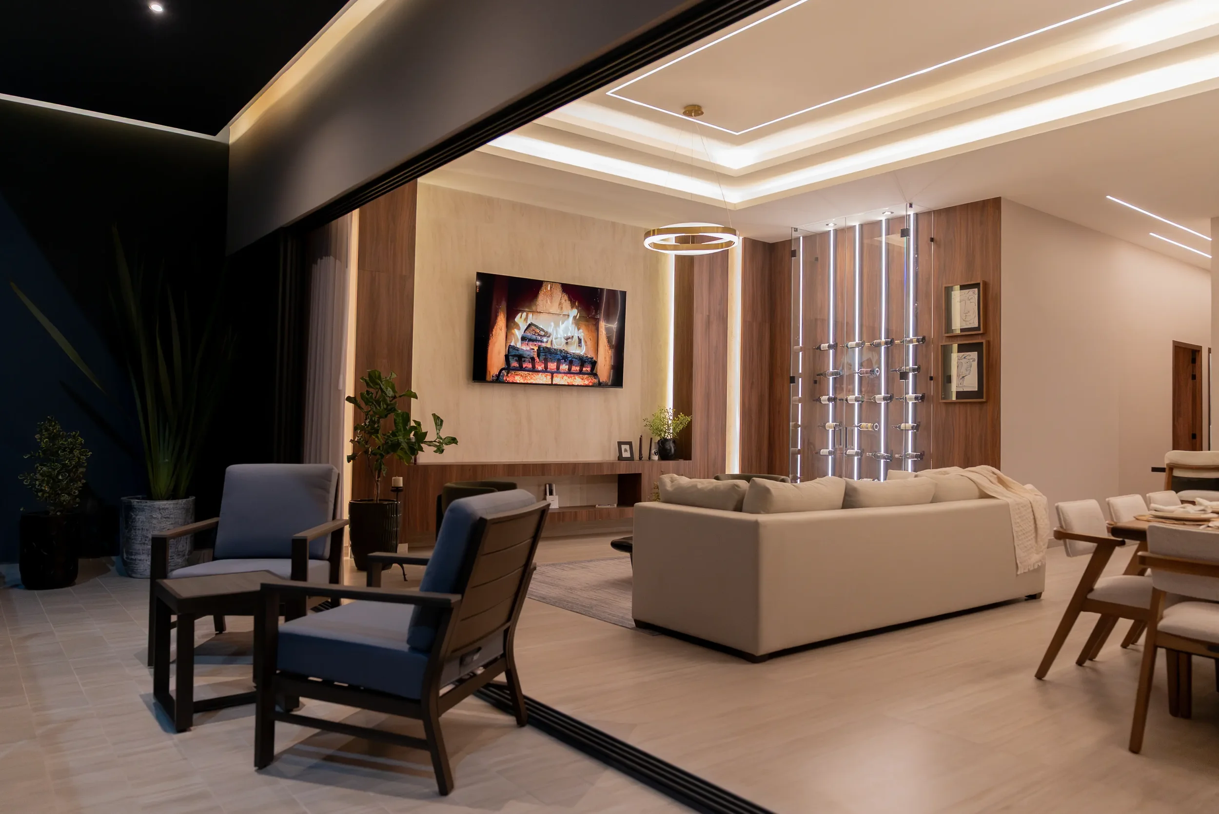 Modern living room with beige sectional sofa, wooden accent walls, wall-mounted TV above fireplace, contemporary chandelier, large windows with vertical blinds, dining table with white chairs, potted plants, and stylish interior decor.