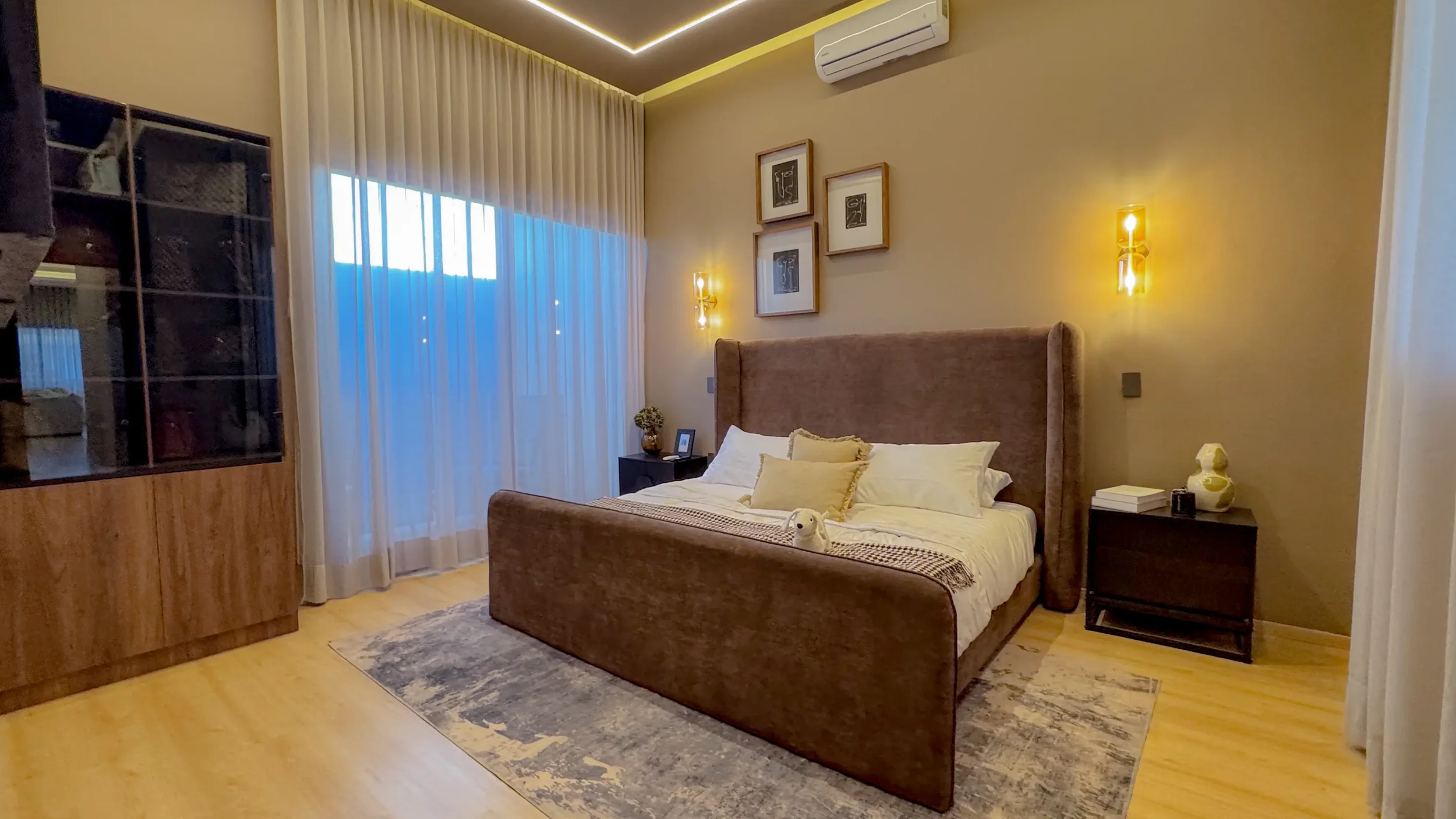 A cozy bedroom with a large bed featuring a beige headboard, surrounded by soft lighting and framed artwork on the wall. There are nightstands with decorative items and books, and a window with sheer curtains allowing natural light to enter.