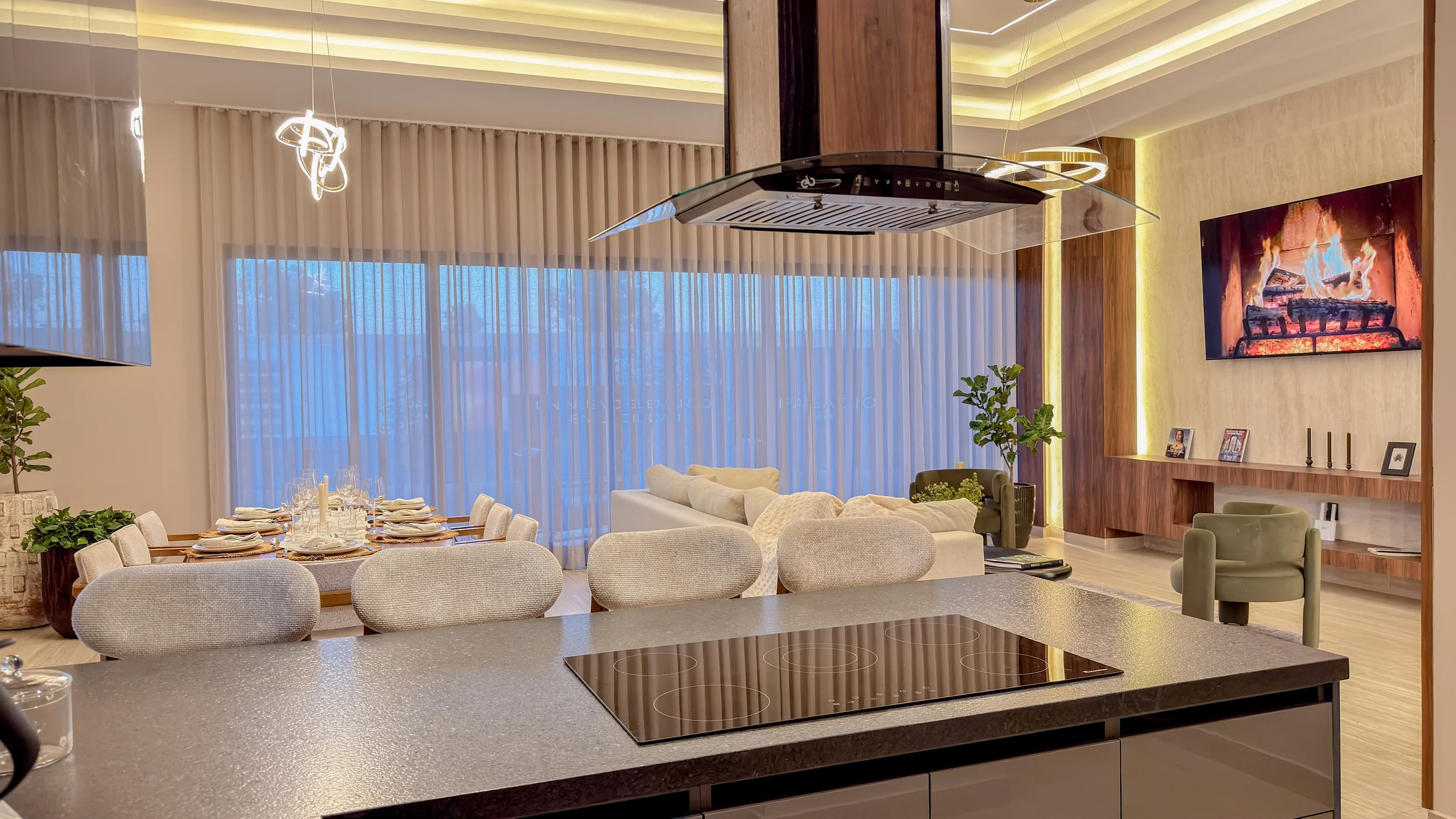 Modern open-concept living and dining area with a kitchen island in the foreground, a dining table set for a meal, a cozy seating area with sofas and armchairs, a large flat-screen TV showing a fireplace, and large windows with sheer curtains letting in natural light.