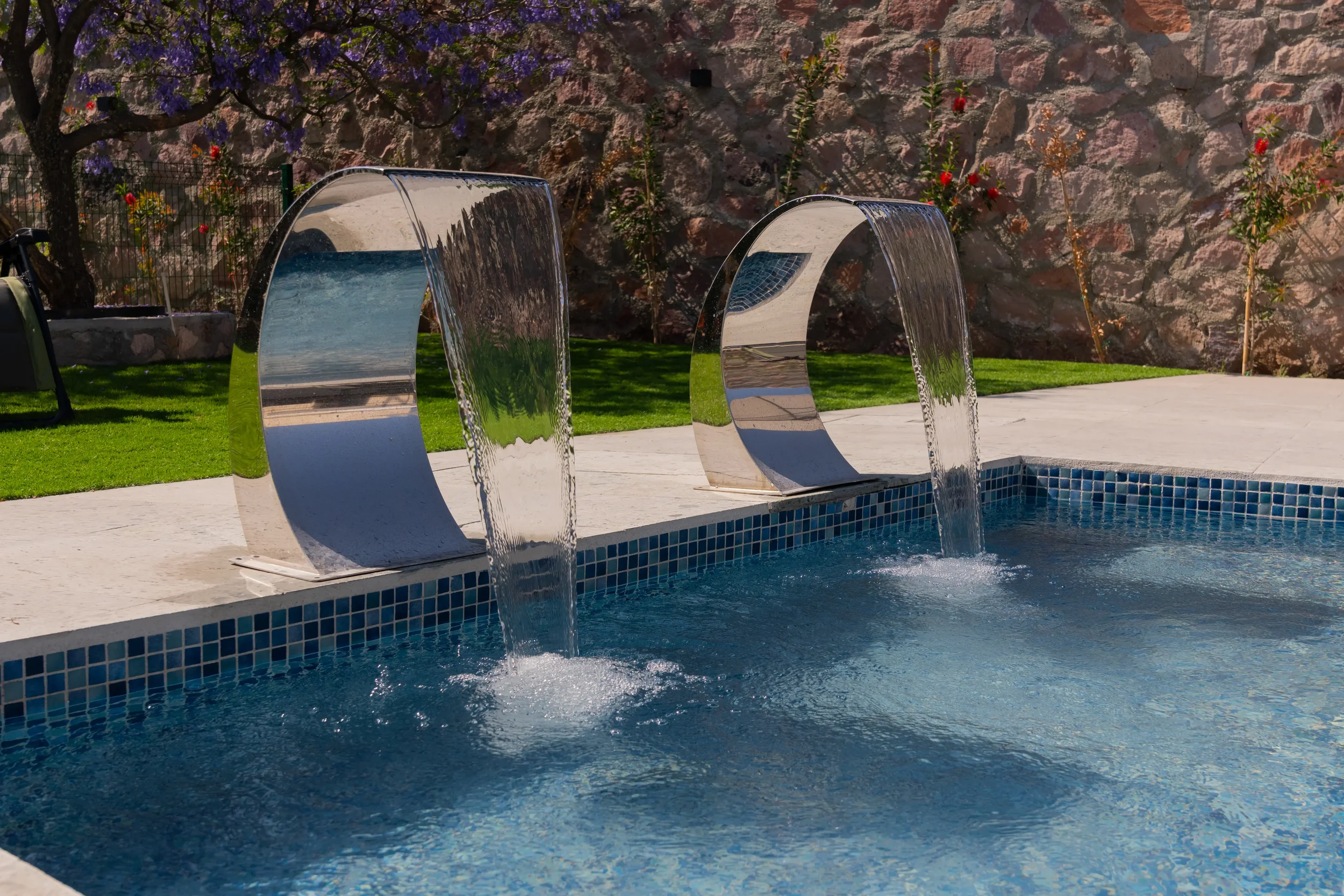 A swimming pool with modern water features pouring water into the pool, surrounded by green grass, flowers, and a stone wall.