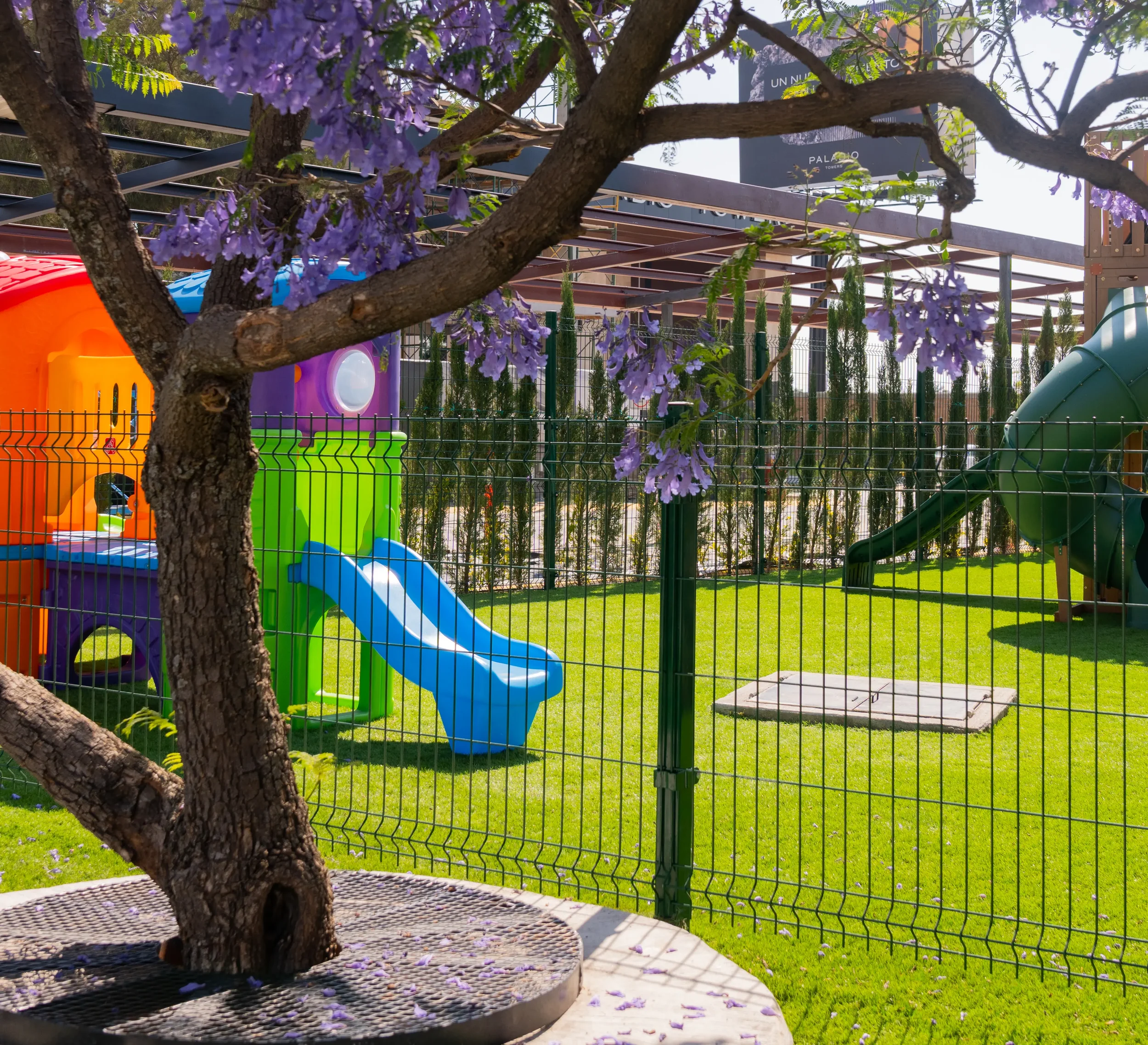 Colorful playground with slides and a shaded tree in a grassy area, enclosed by a black metal fence, with purple flowering tree in the foreground.