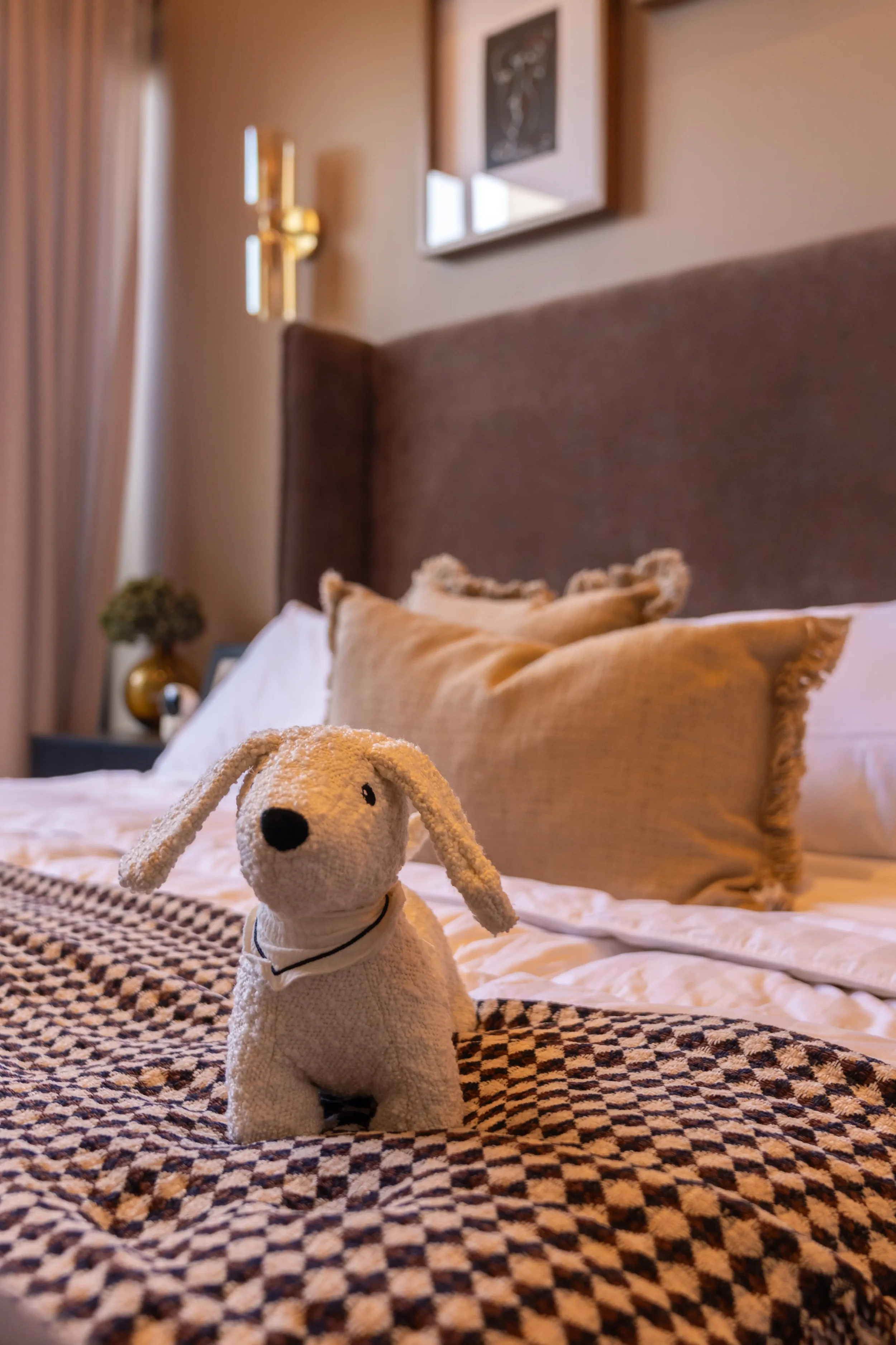 A stuffed plush dog toy with floppy ears, white cloth collar, and black nose positioned on a bed with checkered blanket and beige pillows, in a cozy bedroom setting with framed artwork and gold sconces on the wall.