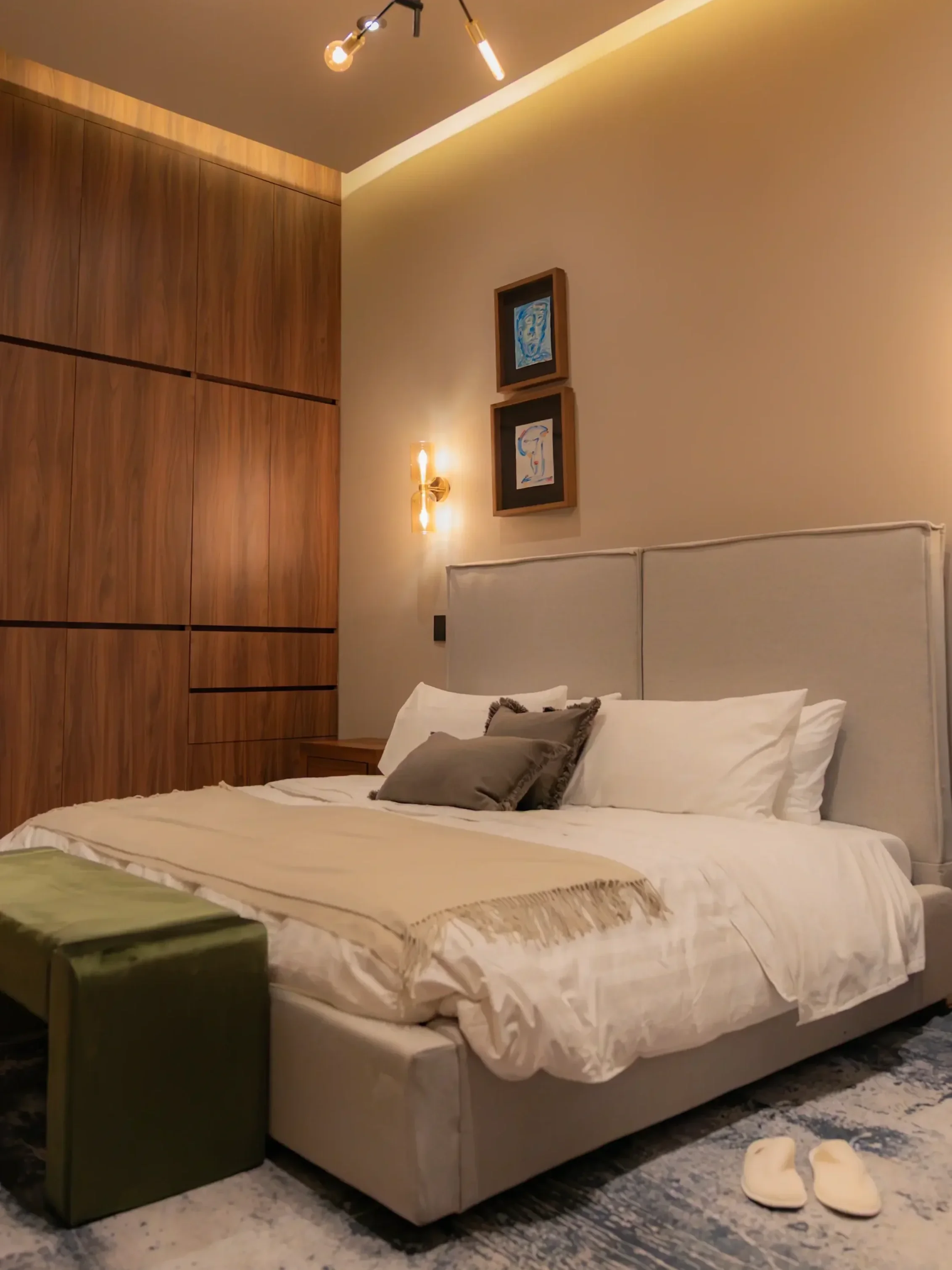 A neatly made bed in a modern bedroom with white linens, gray pillows, a wooden nightstand, wall art, and warm lighting.