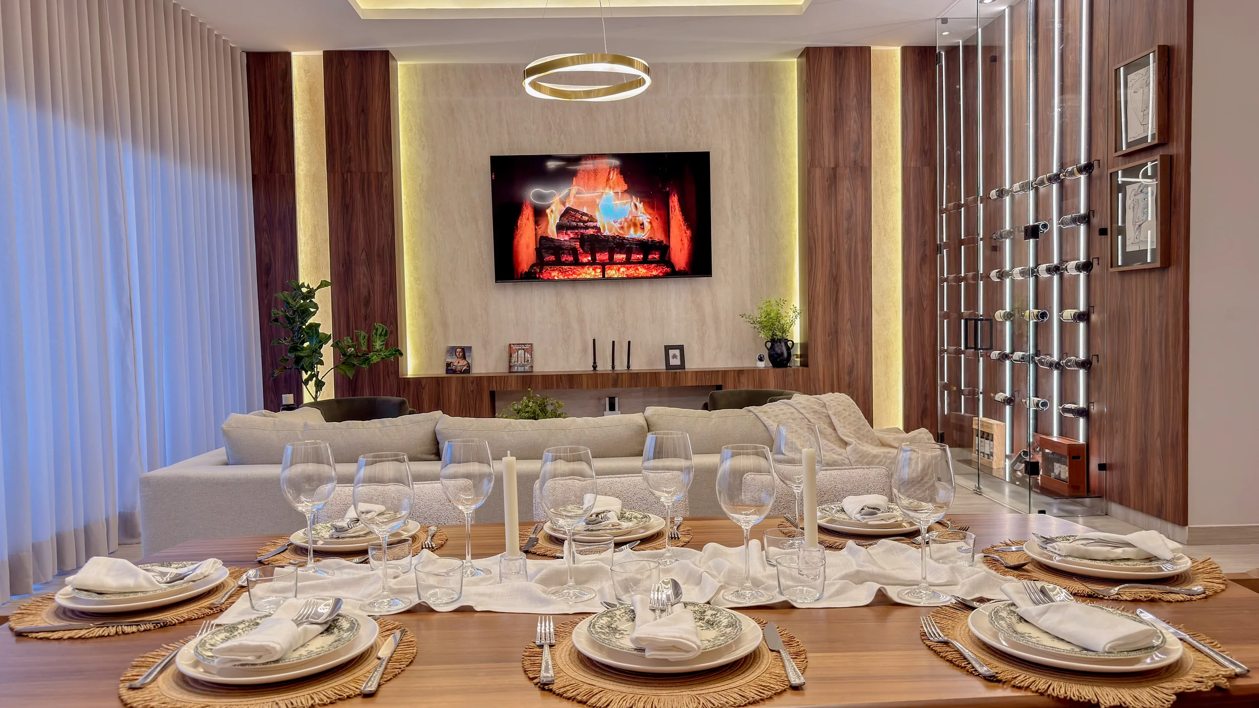 Dining table set for six, decorated with white napkins, glassware, plates, and candles, in an open living room with a TV, cream sofa, plants, and wood paneling.
