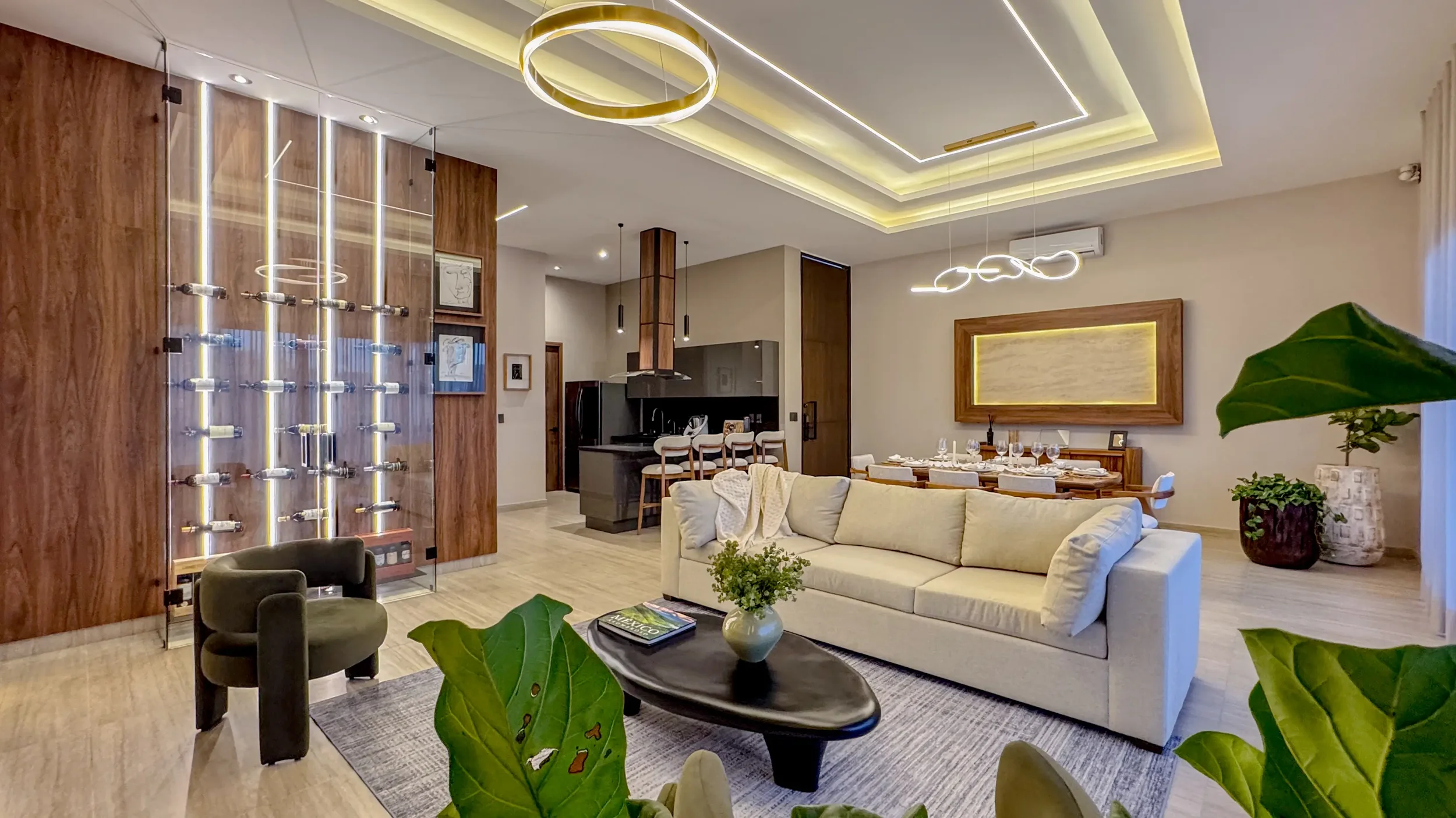 Modern living room with a beige sofa, green plants, a black coffee table with a vase and book, and a wall-mounted wine display with bottles and LED lighting, open kitchen and dining area with a wooden dining table set for a meal, contemporary lighting fixtures, and a ceiling with recessed lighting.