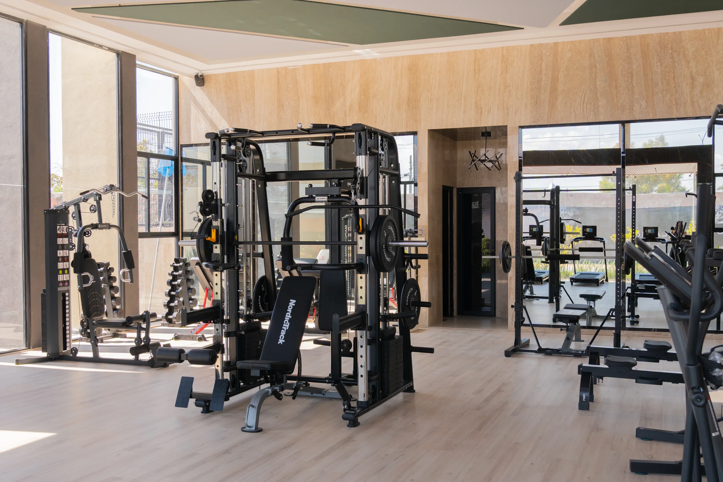 Interior of a modern gym with various workout equipment including a weight machine, dumbbells, and cardio machines, with large windows allowing natural light.