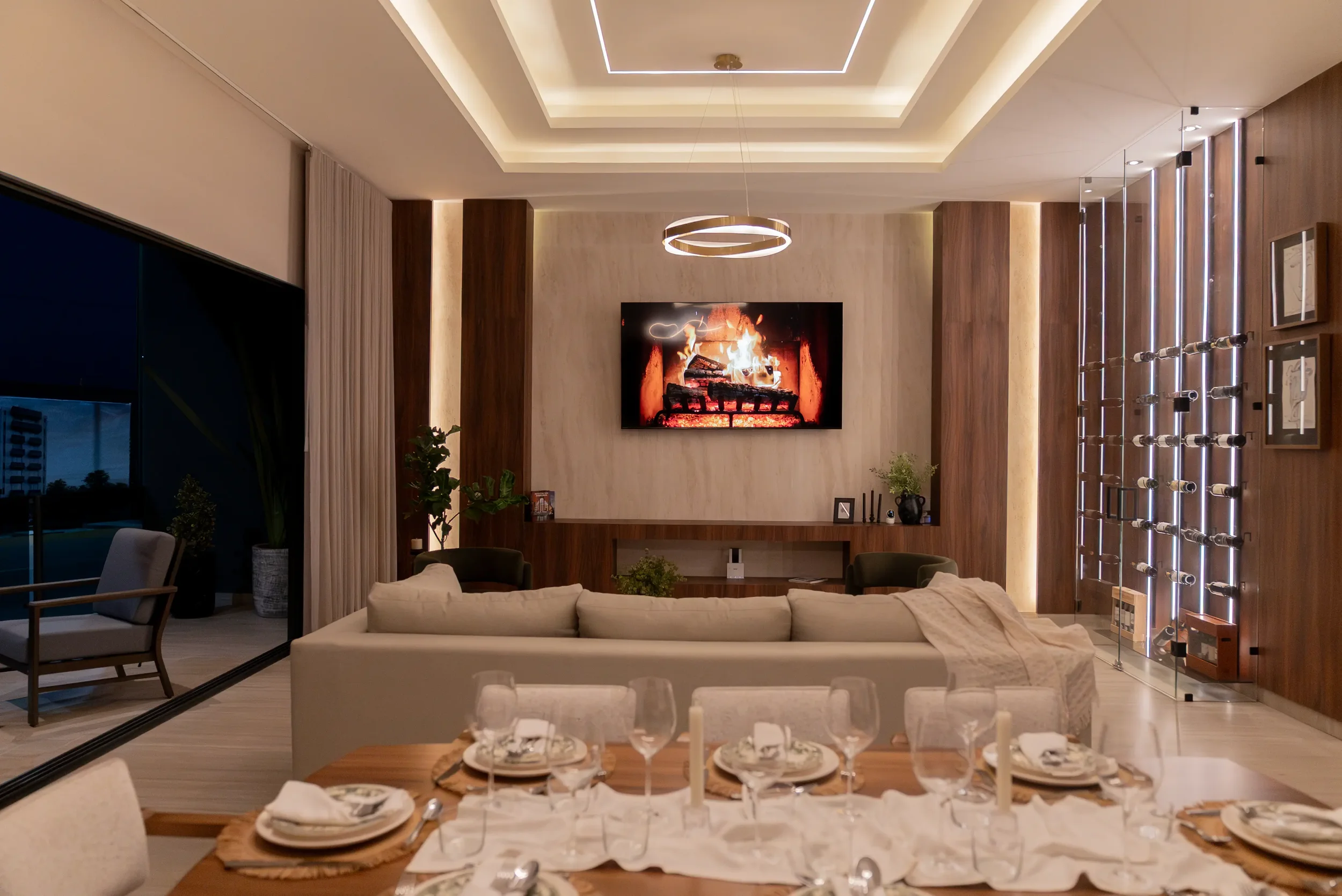 A modern living room with a mounted TV displaying a fireplace scene, a beige sectional sofa, a dining table set with plates, glasses, and cutlery, and large glass doors leading outside. Decorations include plants, framed pictures, and a sculptural wall feature.