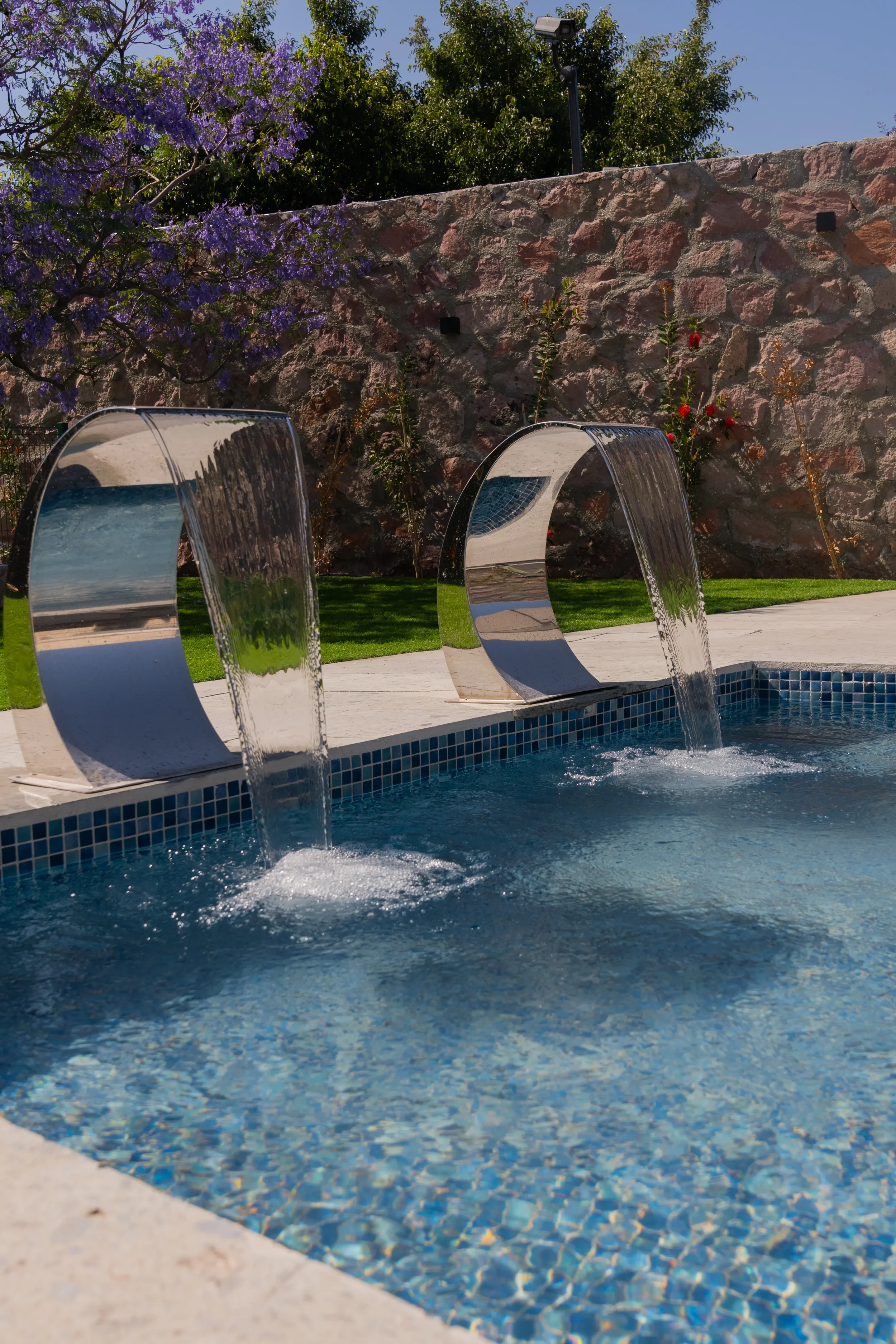 A swimming pool with modern stainless steel waterfalls features a stone wall and greenery in the background.