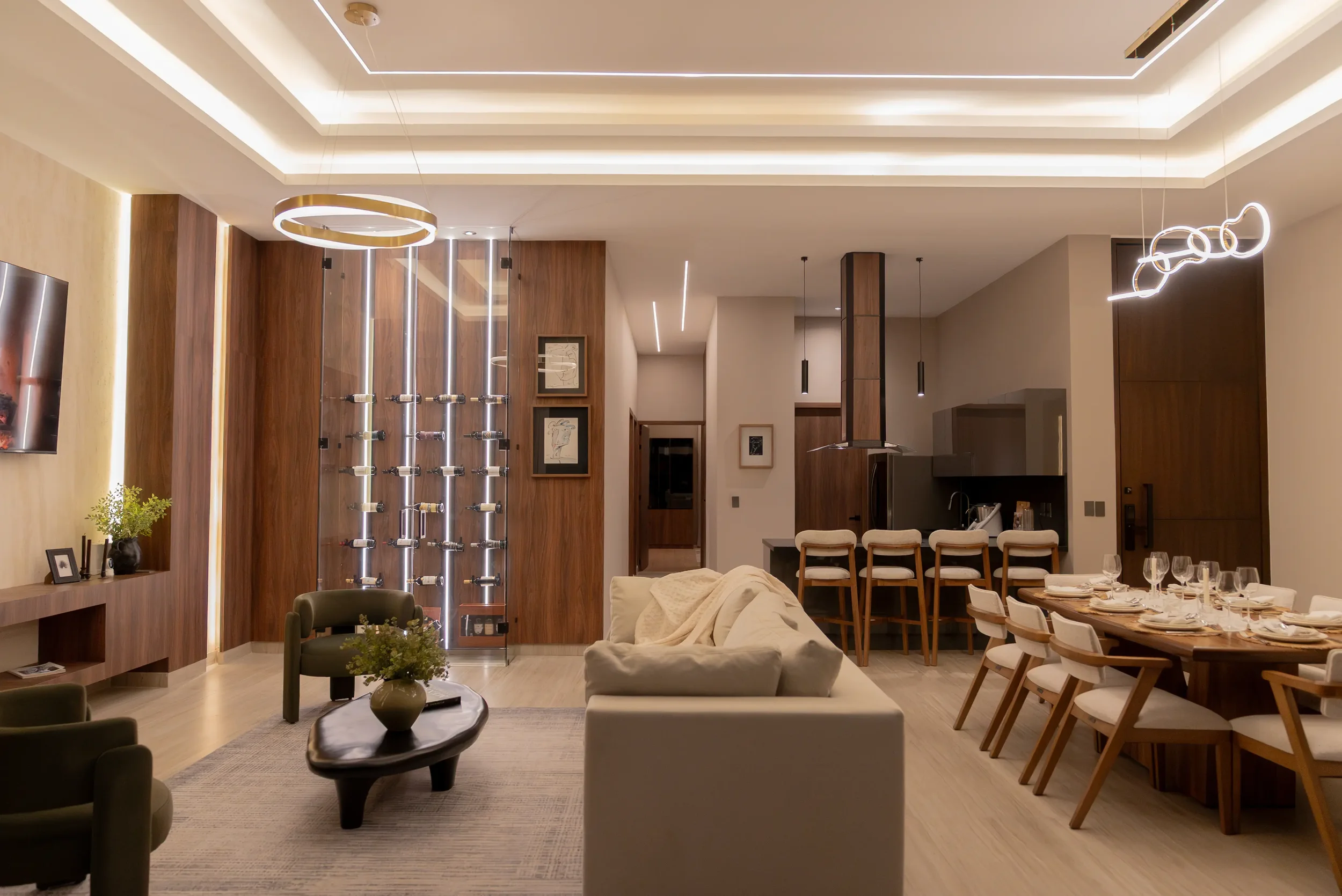 Modern living room and dining area with wooden accents, LED lighting, a beige sofa, a dining table with white chairs, and a wine display.