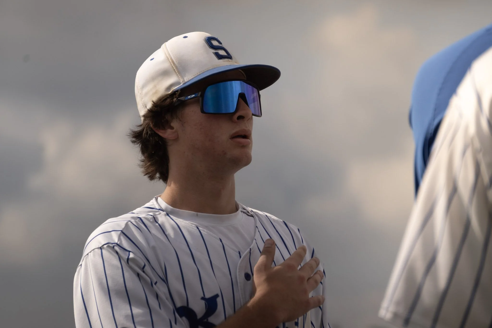 Young man wearing a white baseball cap with a blue 'S', reflective sports sunglasses, and a white and blue striped baseball jersey, touching his chest with his right hand during a national anthem ceremony.