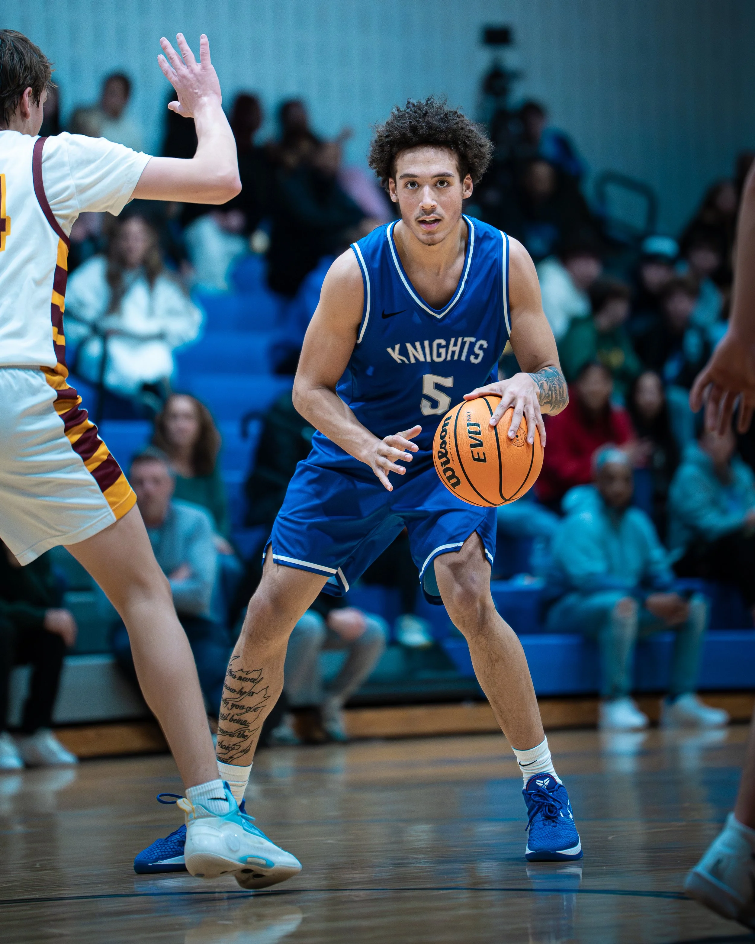 Young male basketball player in a blue uniform with 'Knights' written on it, wearing jersey number 5, dribbling a basketball on indoor court, guarded by opponents, with spectators in the background.