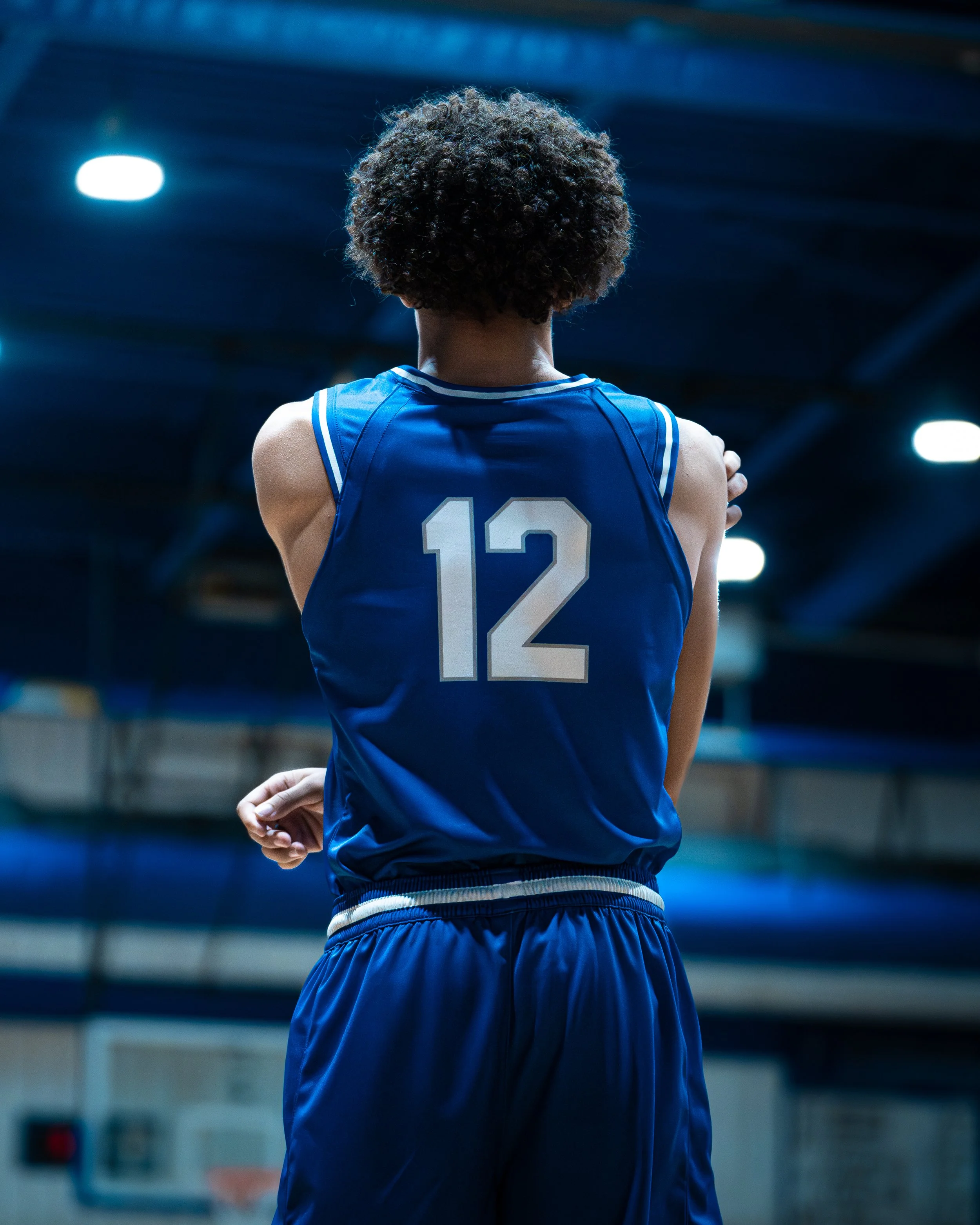 Back view of a female basketball player in a blue jersey with the number 12, standing with arms crossed in an indoor basketball court.