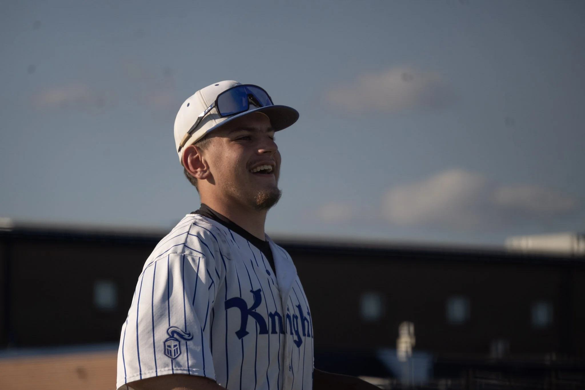 A young man wearing a baseball cap, sunglasses, and a striped baseball jersey, smiling outdoors with a clear sky and a building in the background.