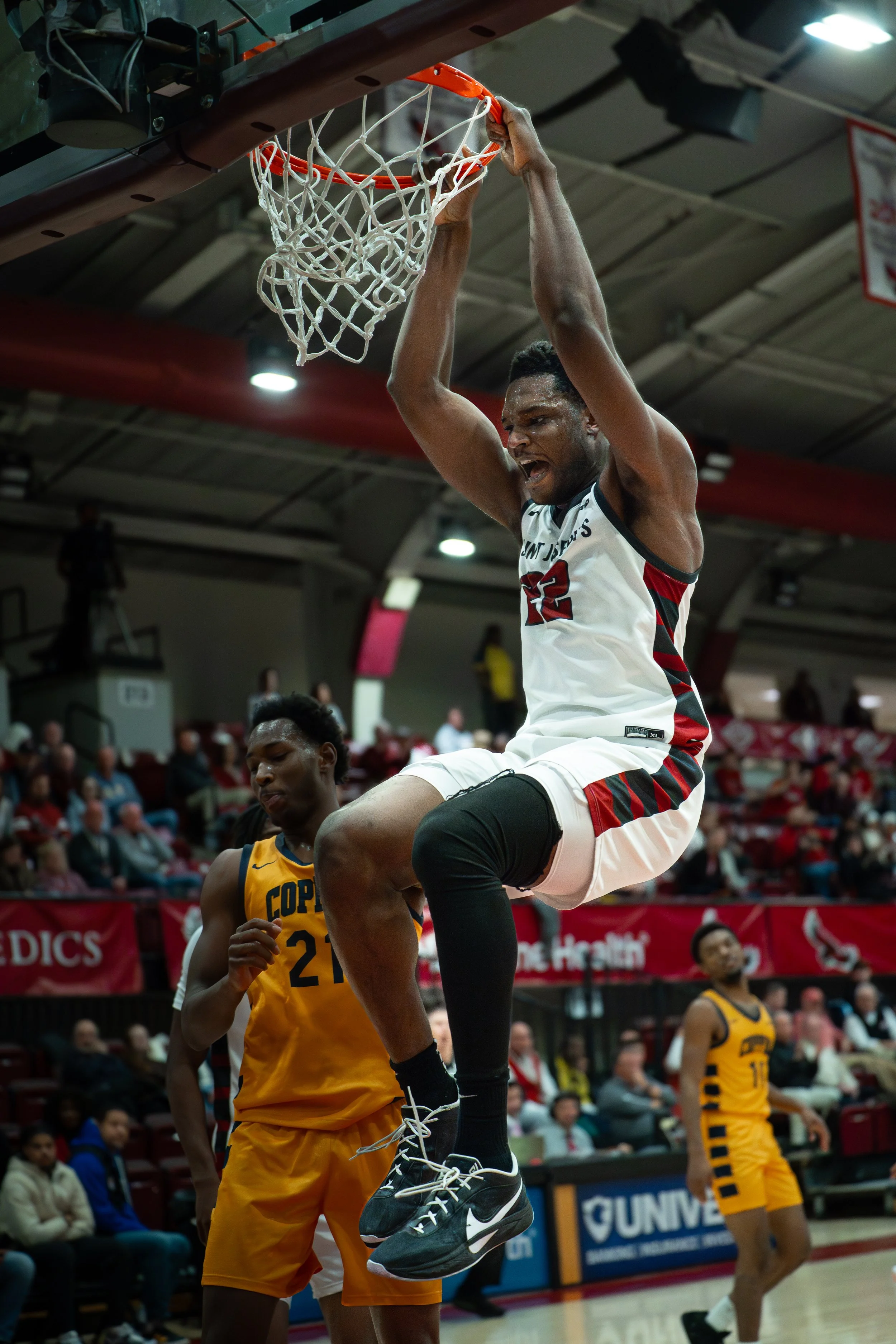 A basketball player wearing a white and red jersey is dunking the ball into the hoop during a game, with another player in a yellow jersey nearby and an audience watching in the background.