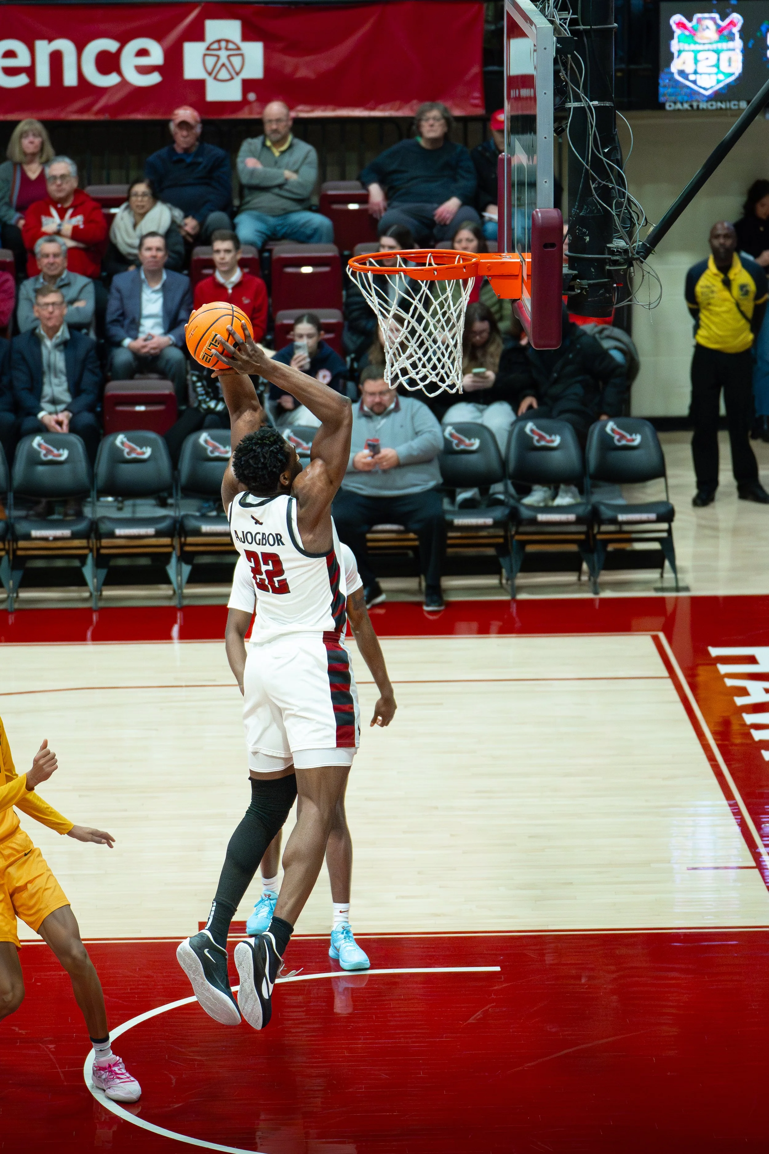 A basketball player jumping to make a shot near the basket, with spectators watching in the background.