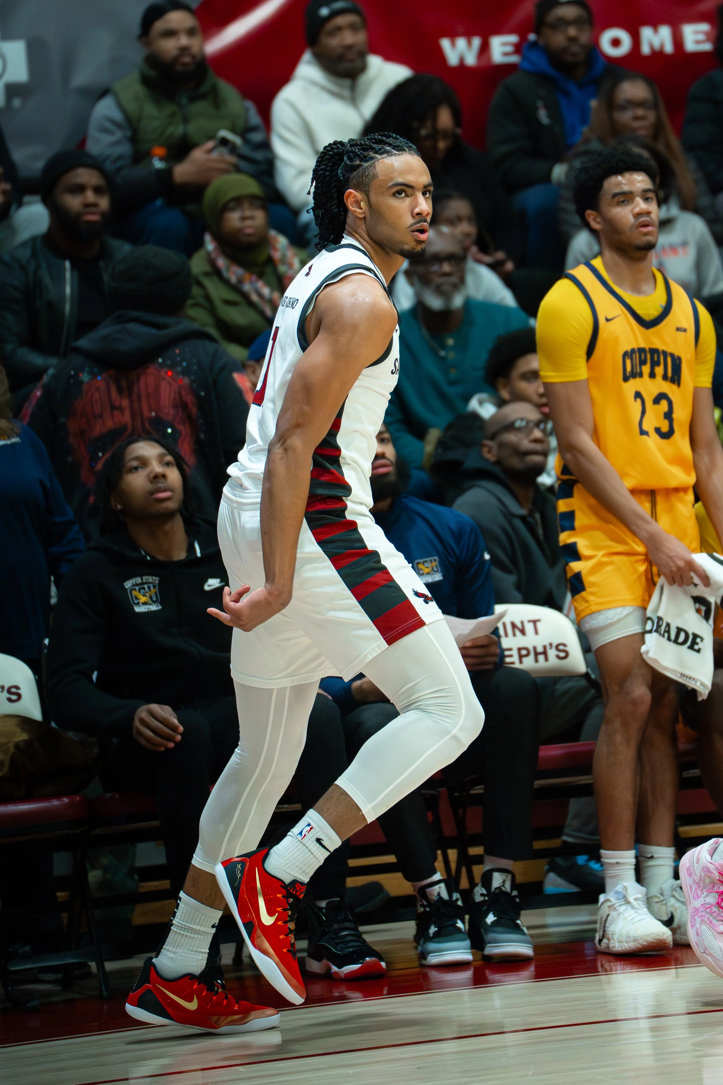A basketball player in white with red and black details, wearing red and yellow Nike sneakers, is in mid-action on the court with a crowd watching in the background.
