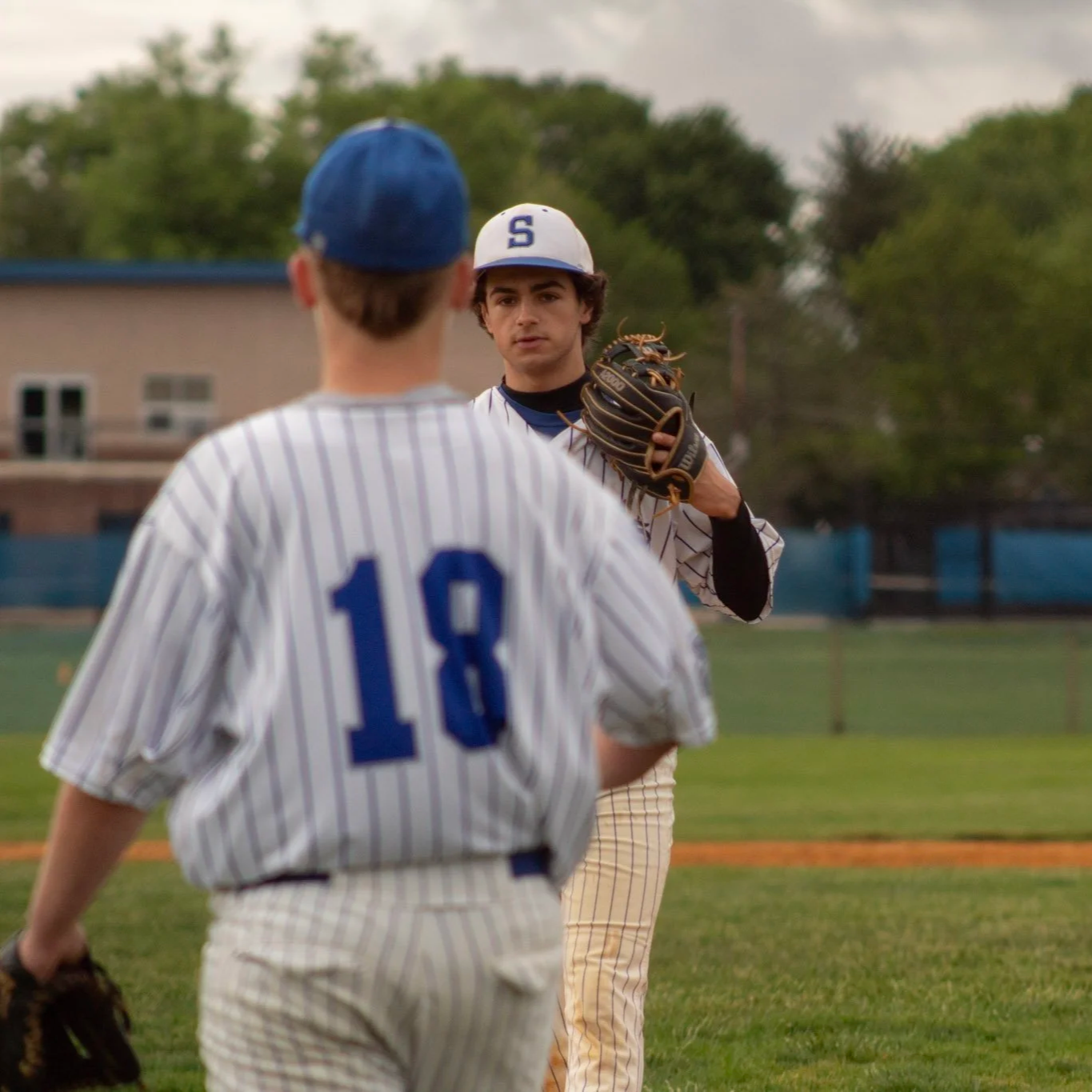 Two young baseball players in uniform on a baseball field during a game or practice. One player, facing away, wears a white jersey with blue pinstripes and the number 18. The other player, facing forward, holds a baseball glove and wears a white jers