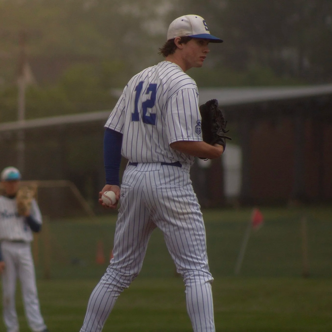 A young male baseball player wearing a white pinstripe uniform with the number 12 on the back, a baseball cap, and a glove on his left hand, holding a baseball in his right hand, standing on a baseball field.