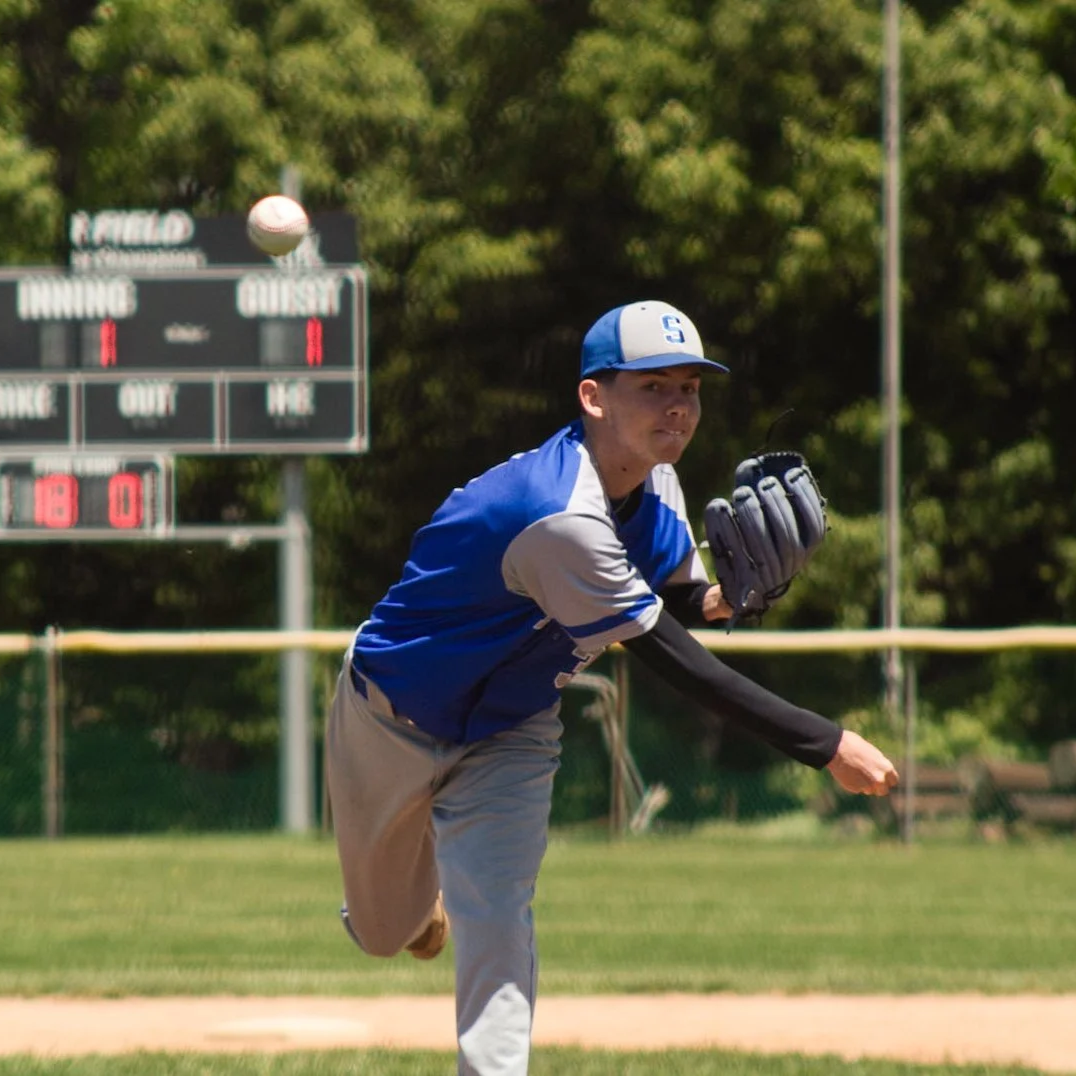 A baseball player in a blue and gray uniform throws a baseball on a field with a scoreboard in the background.