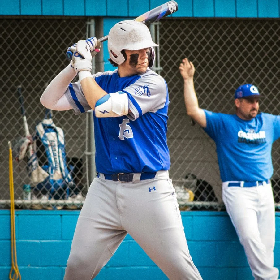 A baseball player wearing a helmet, blue jersey, and white pants, prepares to swing a bat, with a coach or teammate in the background near a chain-link fence.