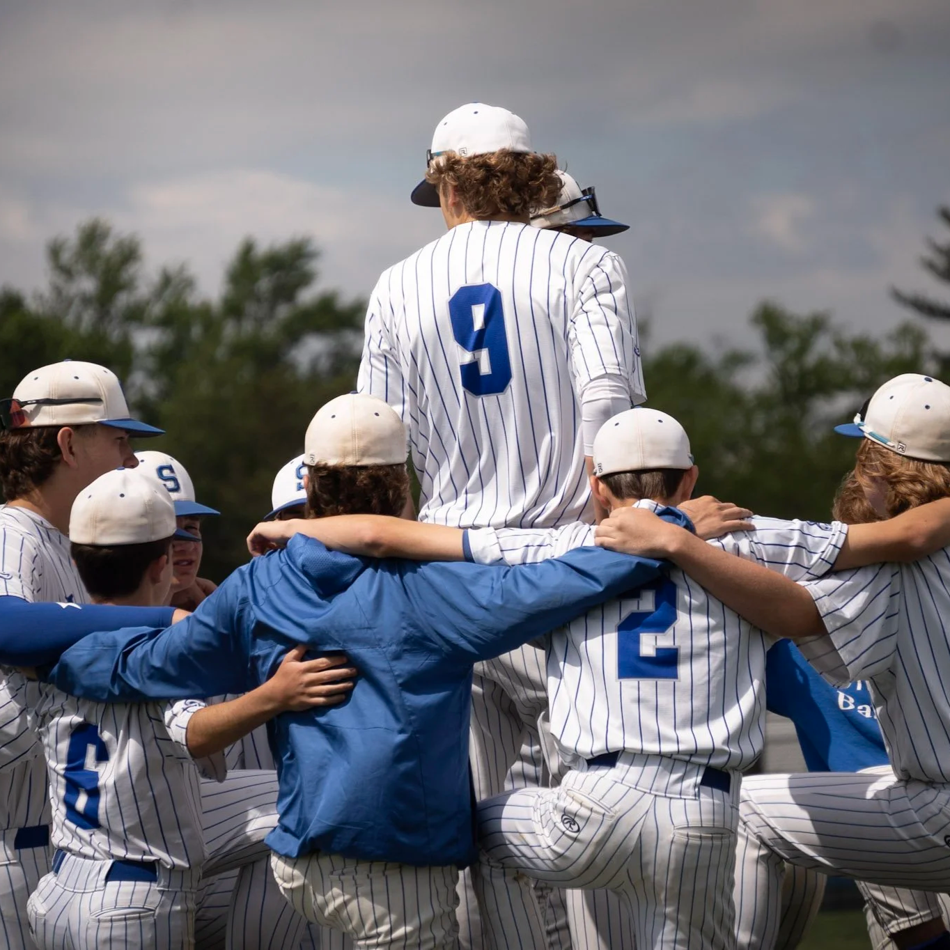 A group of baseball players wearing striped uniforms and caps, celebrating a victory outdoors, with one player being lifted by teammates.