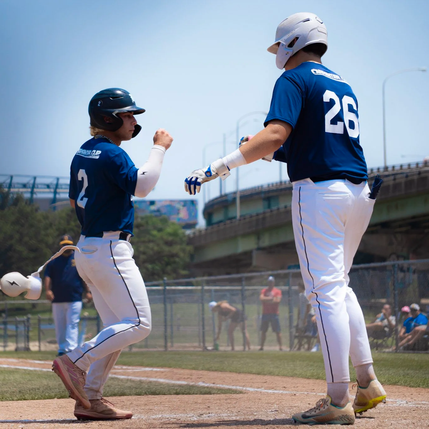 Two baseball players in blue jerseys and white pants are on the field; one player with a black helmet is running towards the other player who is wearing a white helmet, possibly celebrating during a game on a sunny day.