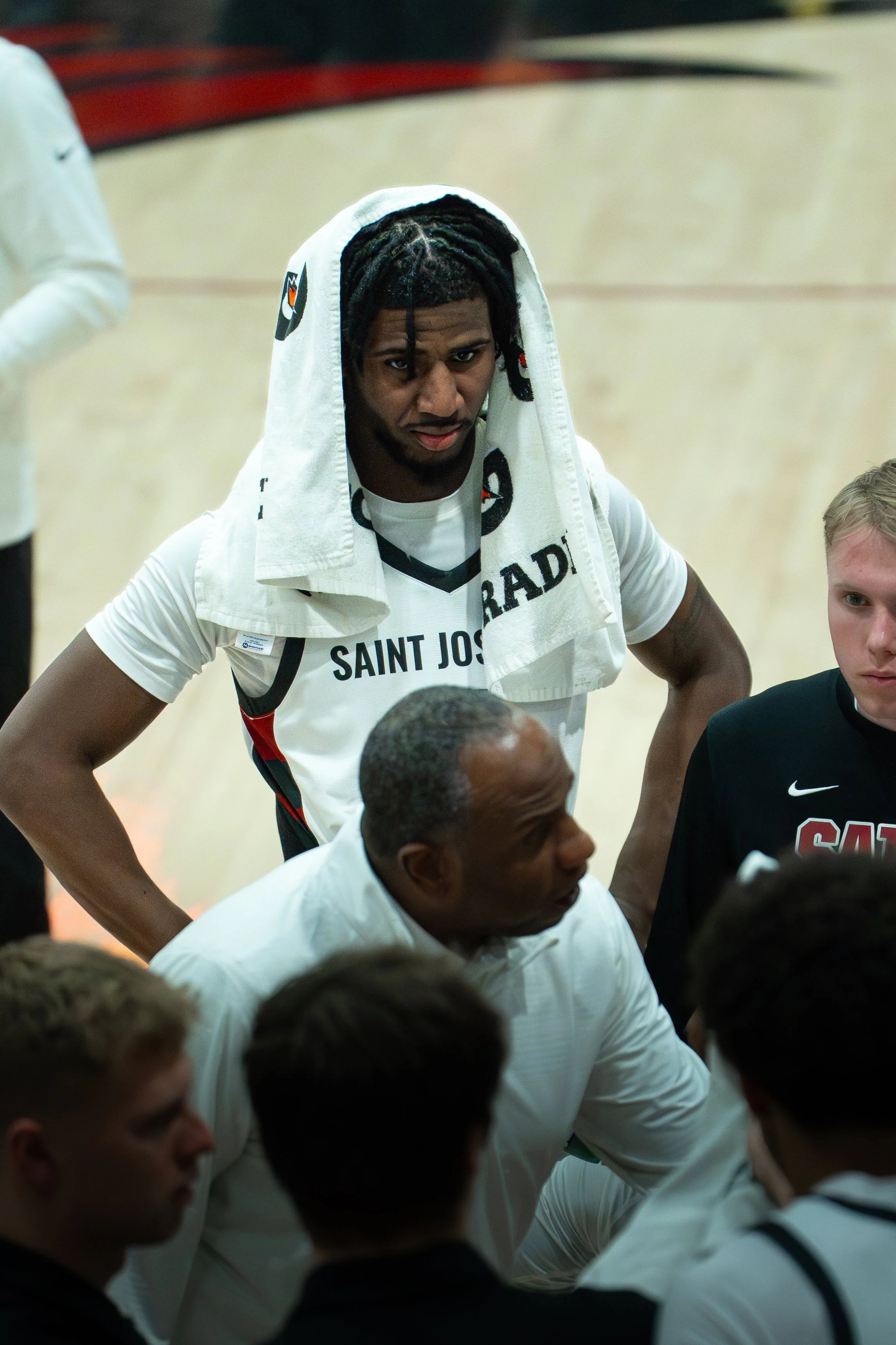 A young African American man with dreadlocks and a towel over his head, wearing a "Saint Joseph" basketball jersey, standing with his hands on his hips during a team discussion.
