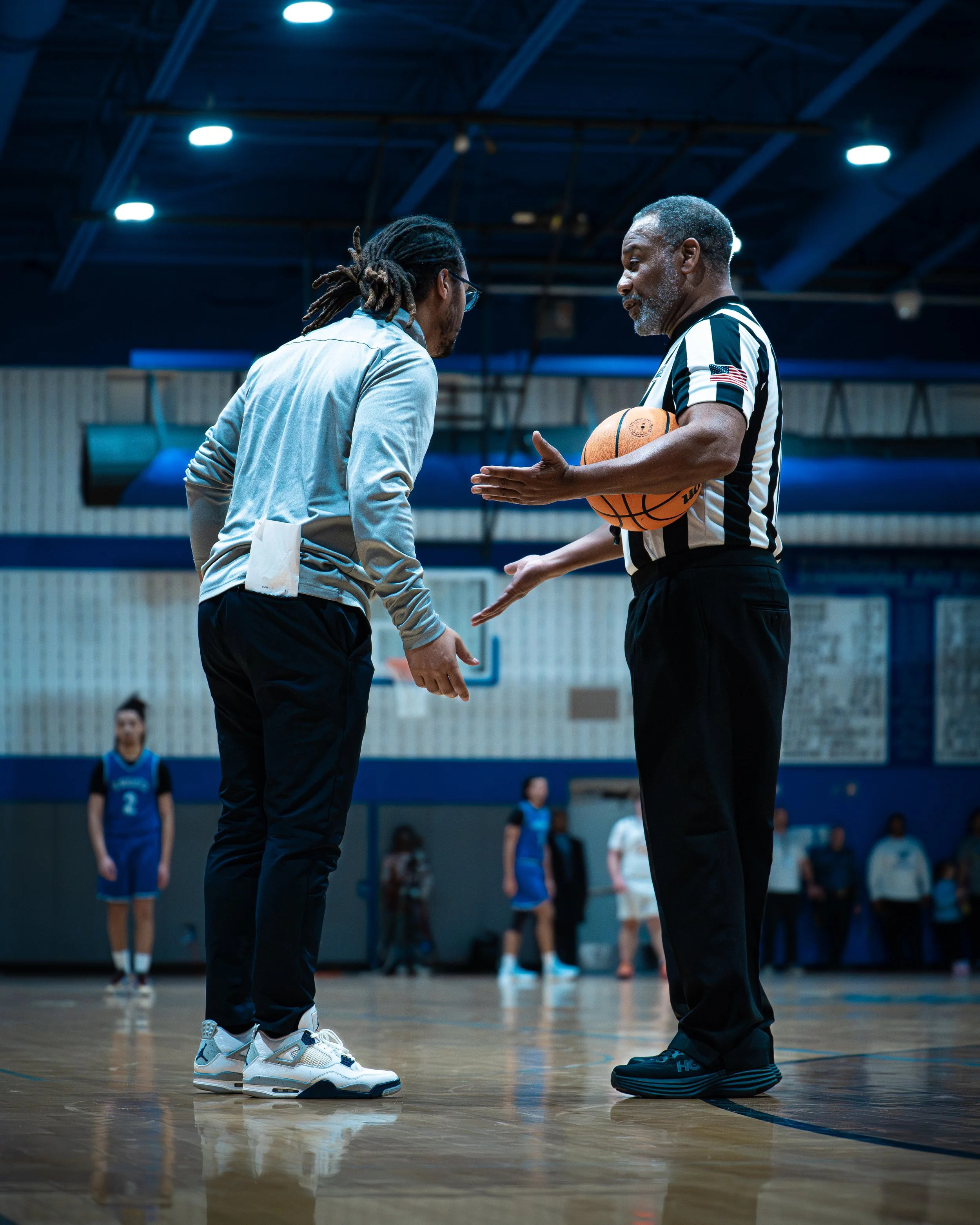 A basketball referee holding a basketball explains a call to a young player on an indoor basketball court. Other players and spectators are visible in the background.